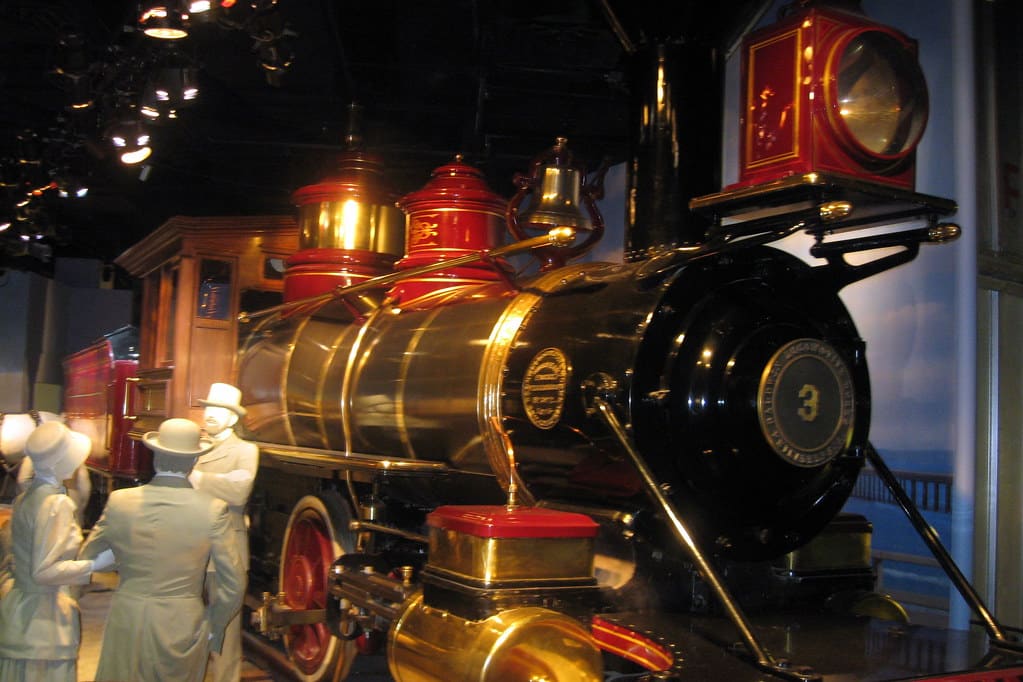 A shiny, black and gold vintage steam locomotive with the number 3 on its front is displayed indoors at one of the Best Smithsonian Museums for Student Groups. Mannequins dressed in white period clothing stand nearby, appearing to observe the train.