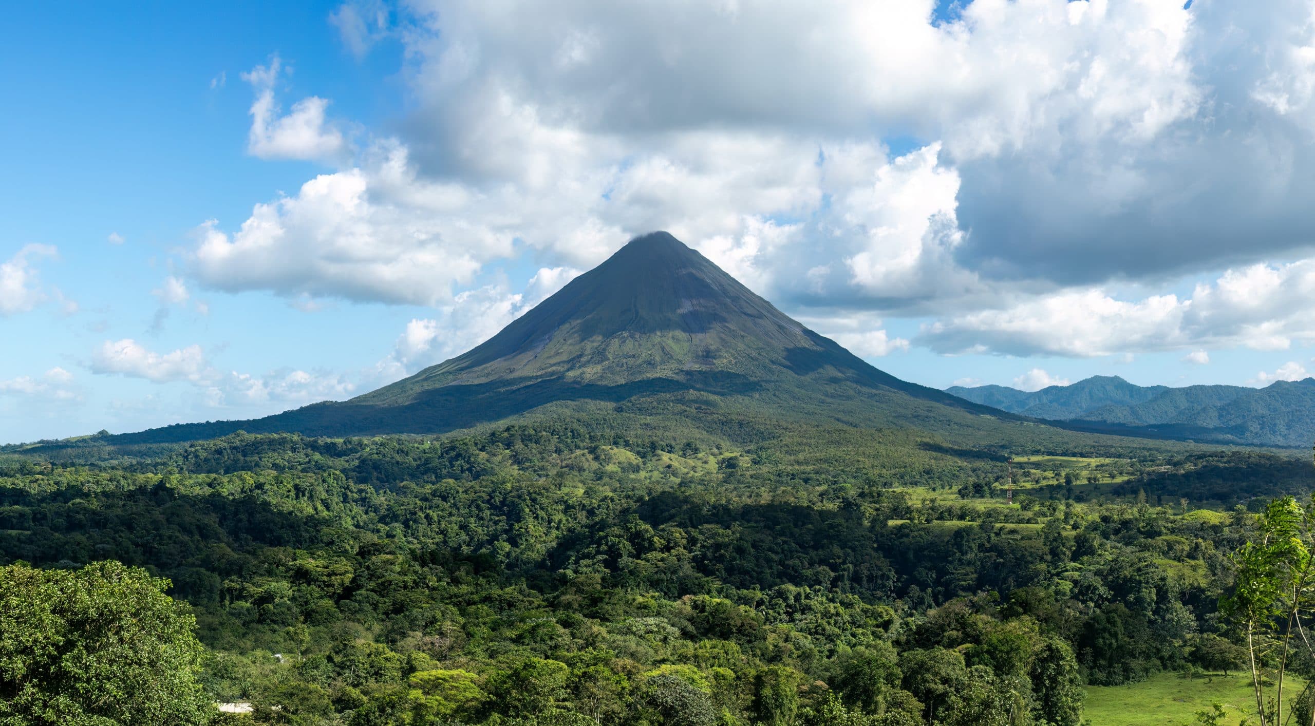 A cone-shaped volcano rises above a lush, green forest under a partly cloudy sky, with mountains visible in the distance.