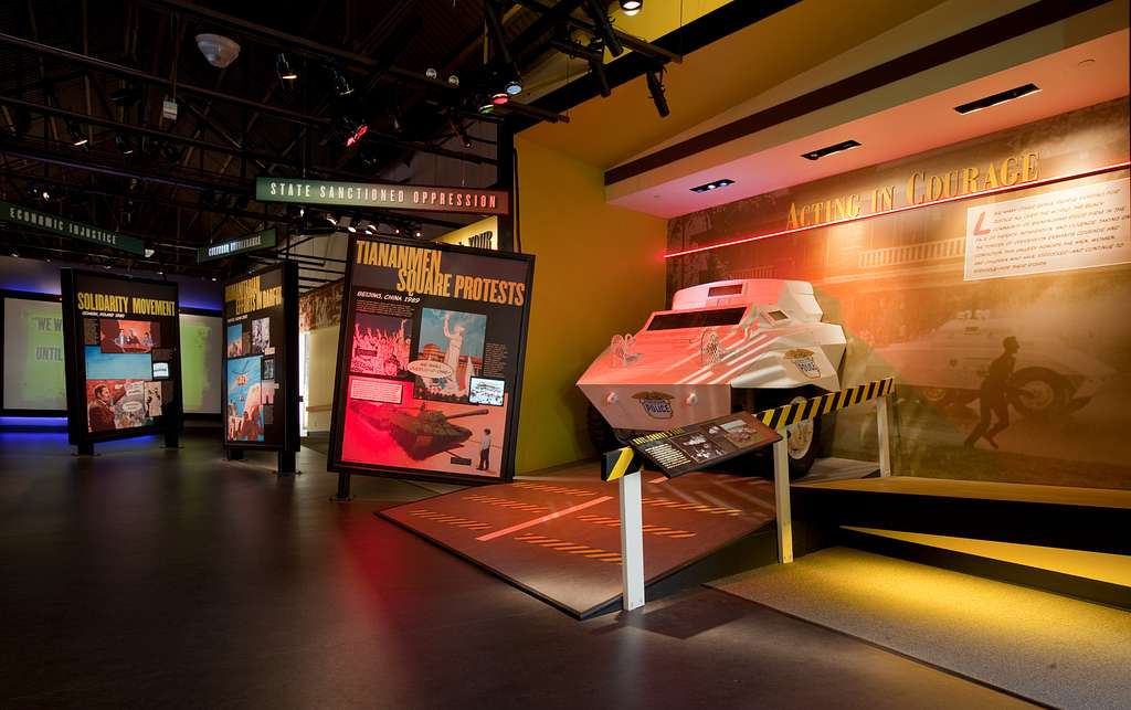 A museum exhibit features a white armored vehicle, large panels about the Tiananmen Square protests, and displays on state oppression and acts of courage, with informative text and images under warm lighting.