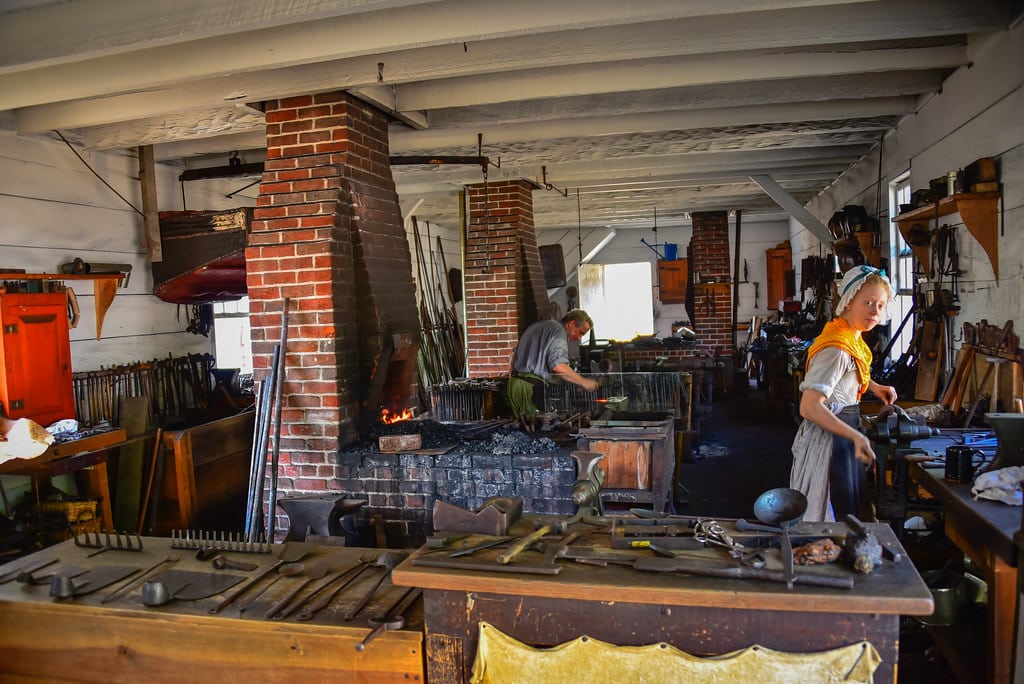 A historical blacksmith workshop during a Williamsburg School Trip, with two people at work—one tending to a fire at the brick forge, the other in a yellow scarf working at a table surrounded by tools and equipment.