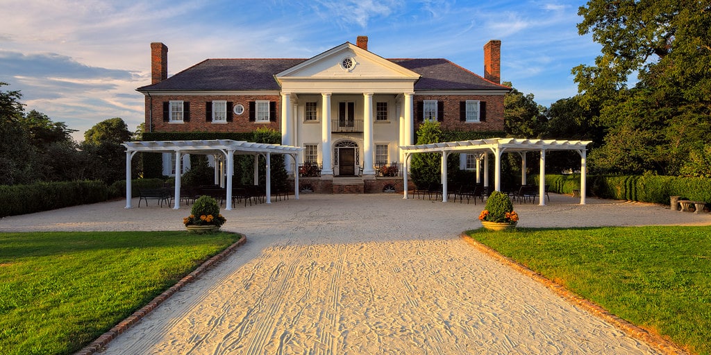 A large, two-story brick mansion with white columns and a covered entrance, flanked by two white pergolas along a wide sandy pathway, surrounded by manicured lawns and trees under a blue sky—perfect for Charleston Field Trips.