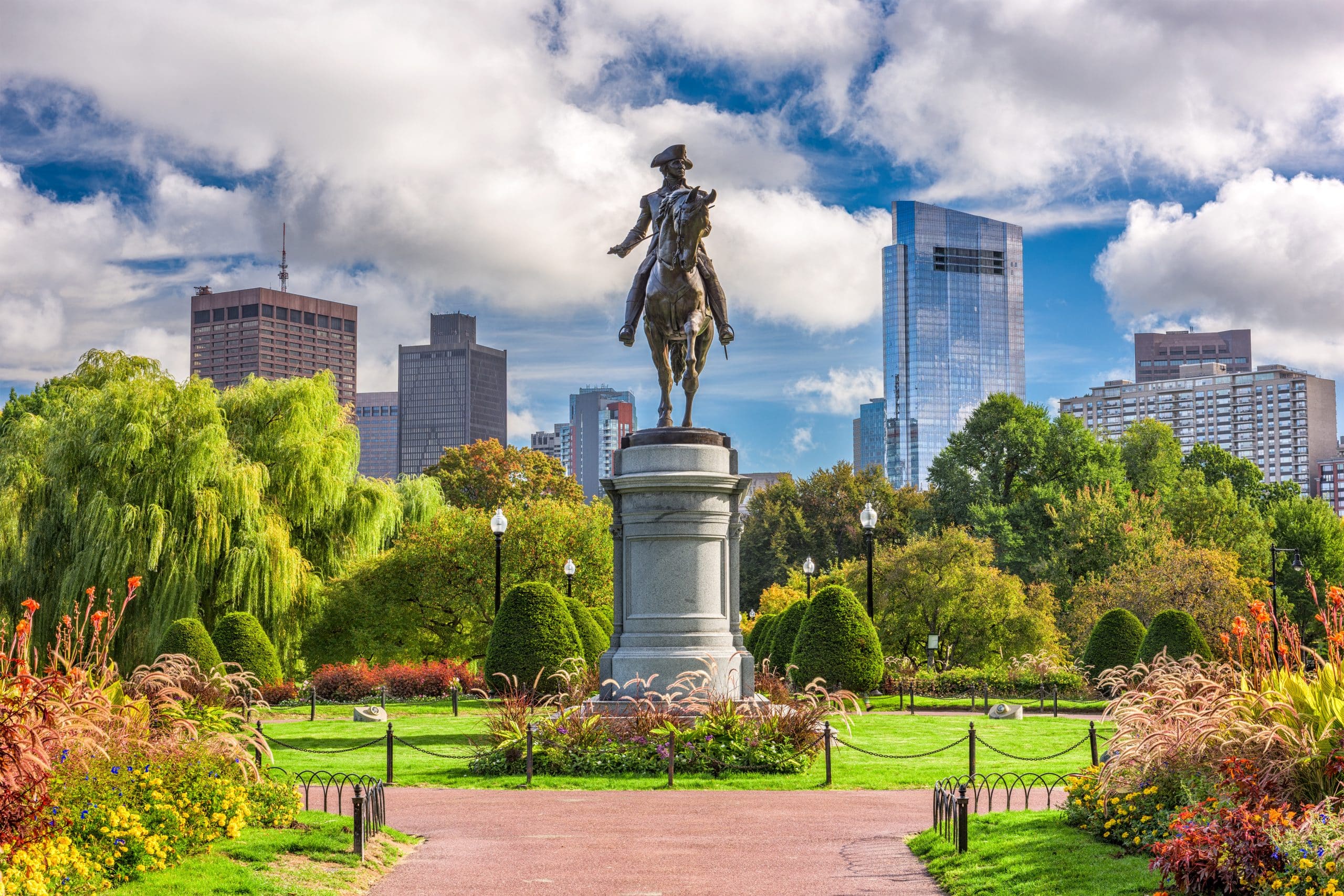 A bronze equestrian statue on a pedestal stands in a lush city park, surrounded by trees and colorful flowers—an iconic sight for any School Trip to Boston—with tall modern buildings under a partly cloudy sky in the background.