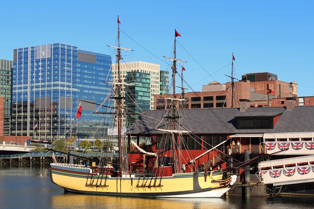 A historic tall ship with yellow sides is docked near a red building decorated with bunting, with modern glass and brick office buildings in the background under a clear blue sky.