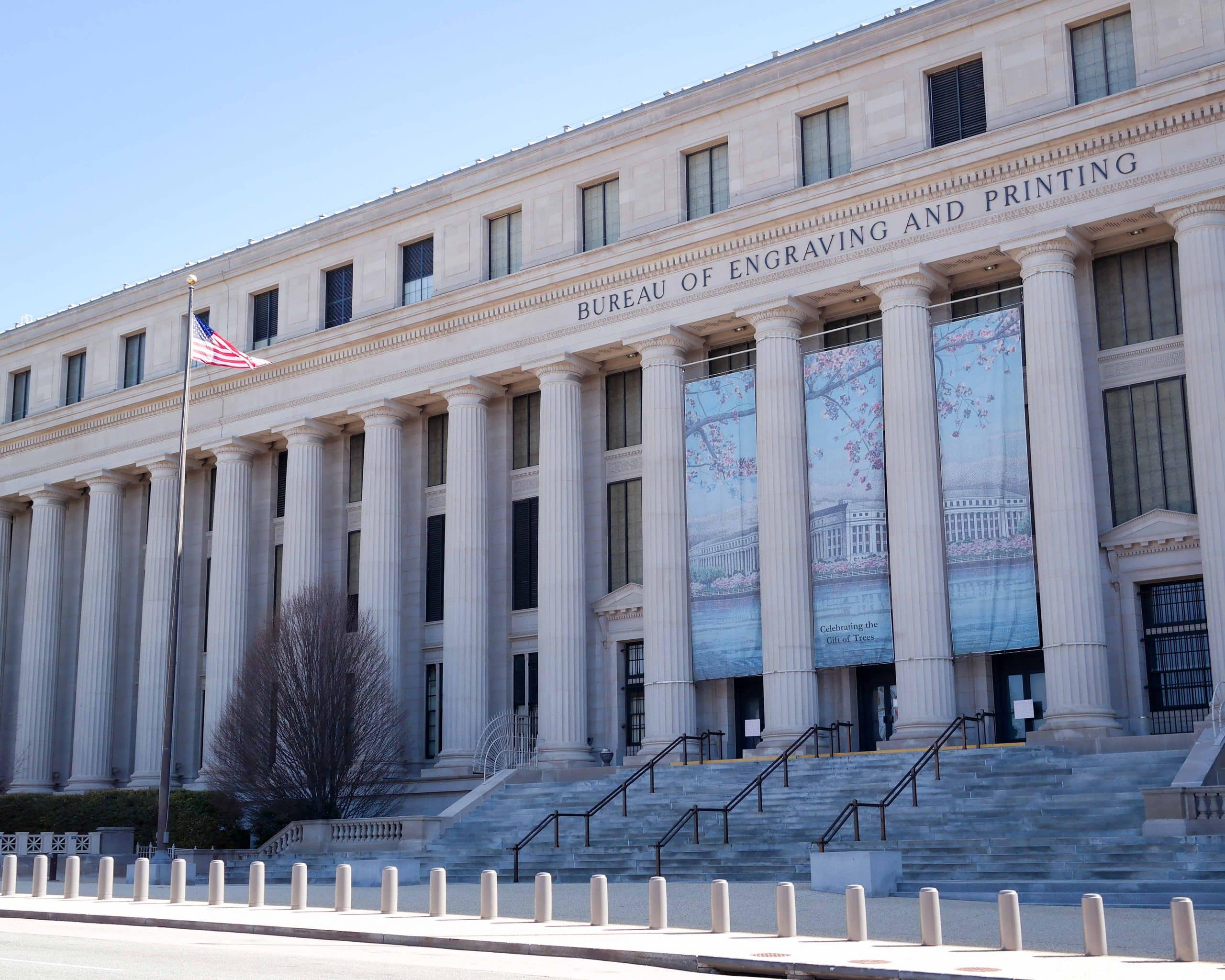 A large neoclassical building with tall columns labeled "Bureau of Engraving and Printing." An American flag flies on a pole, and banners with artwork hang above the entrance—an inspiring stop for STEM Washington DC school trip ideas.