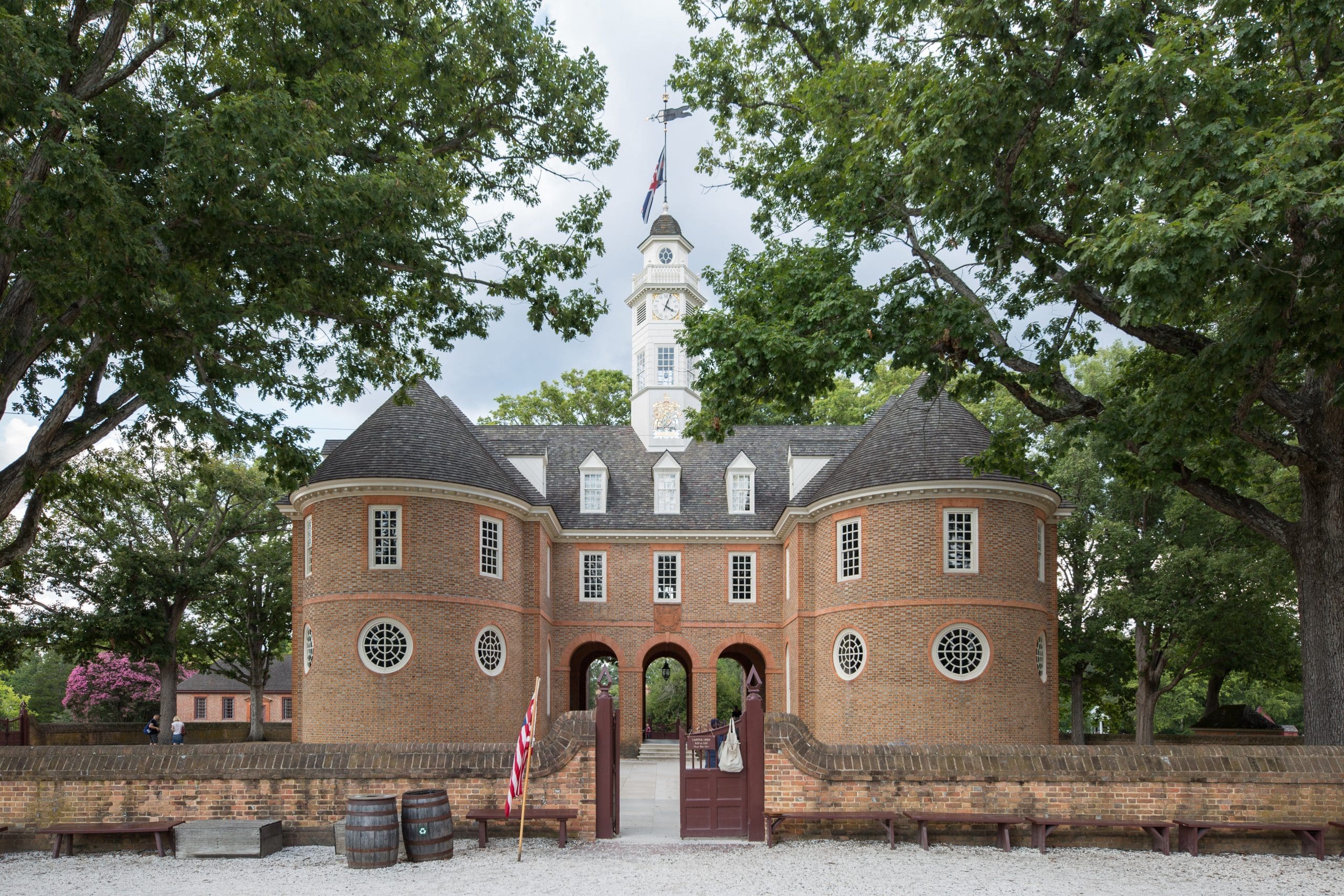 A historic brick building with a central clock tower, arched entryway, and symmetrical wings—reminiscent of a Colonial Williamsburg field trip—stands among large trees and a brick wall, with American flags and barrels near the entrance.