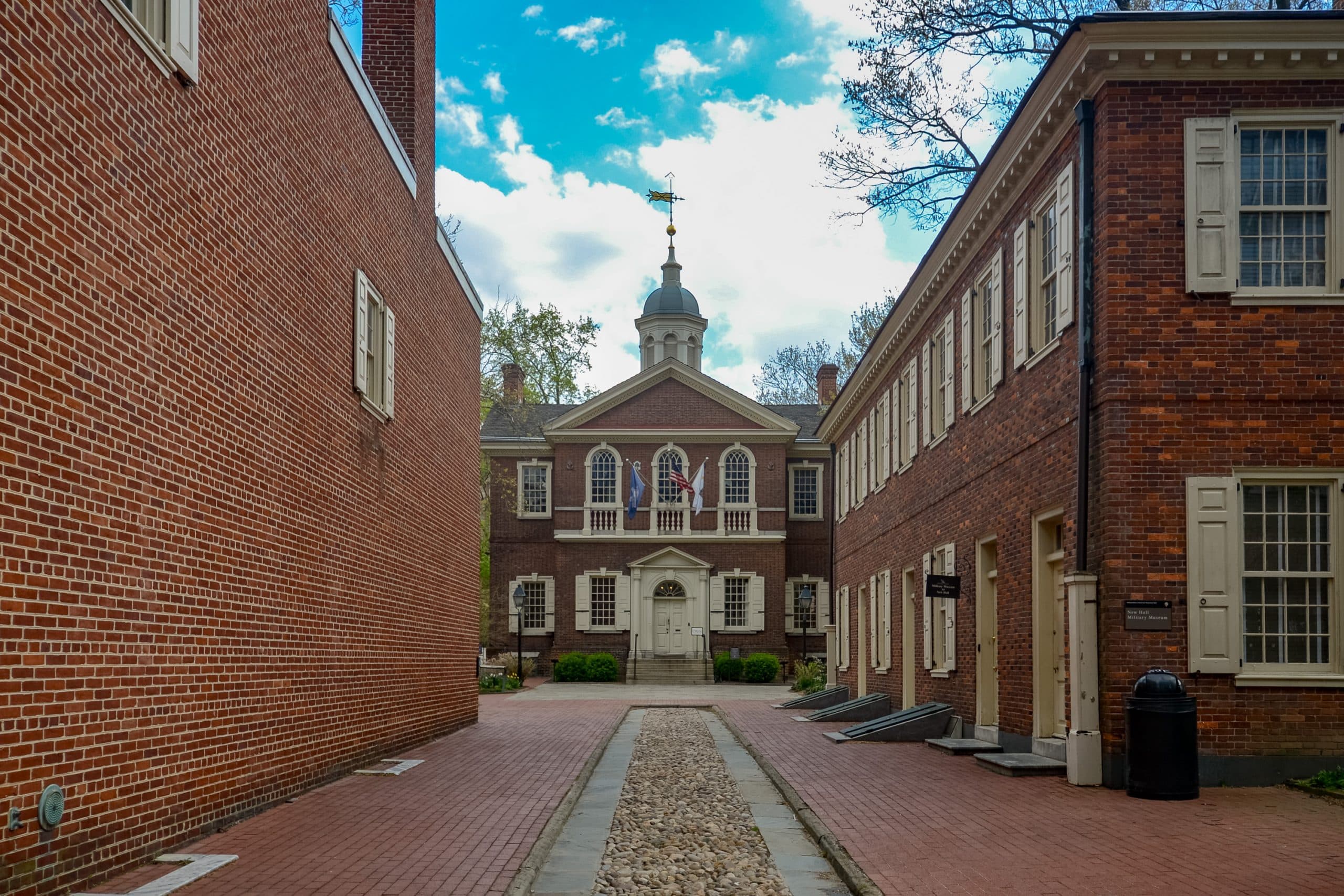 A view of Independence Hall in Philadelphia, one of the top Philadelphia Historical Sites for Students, framed by two red-brick buildings, with a cloudy blue sky above and a brick pathway leading to the entrance.