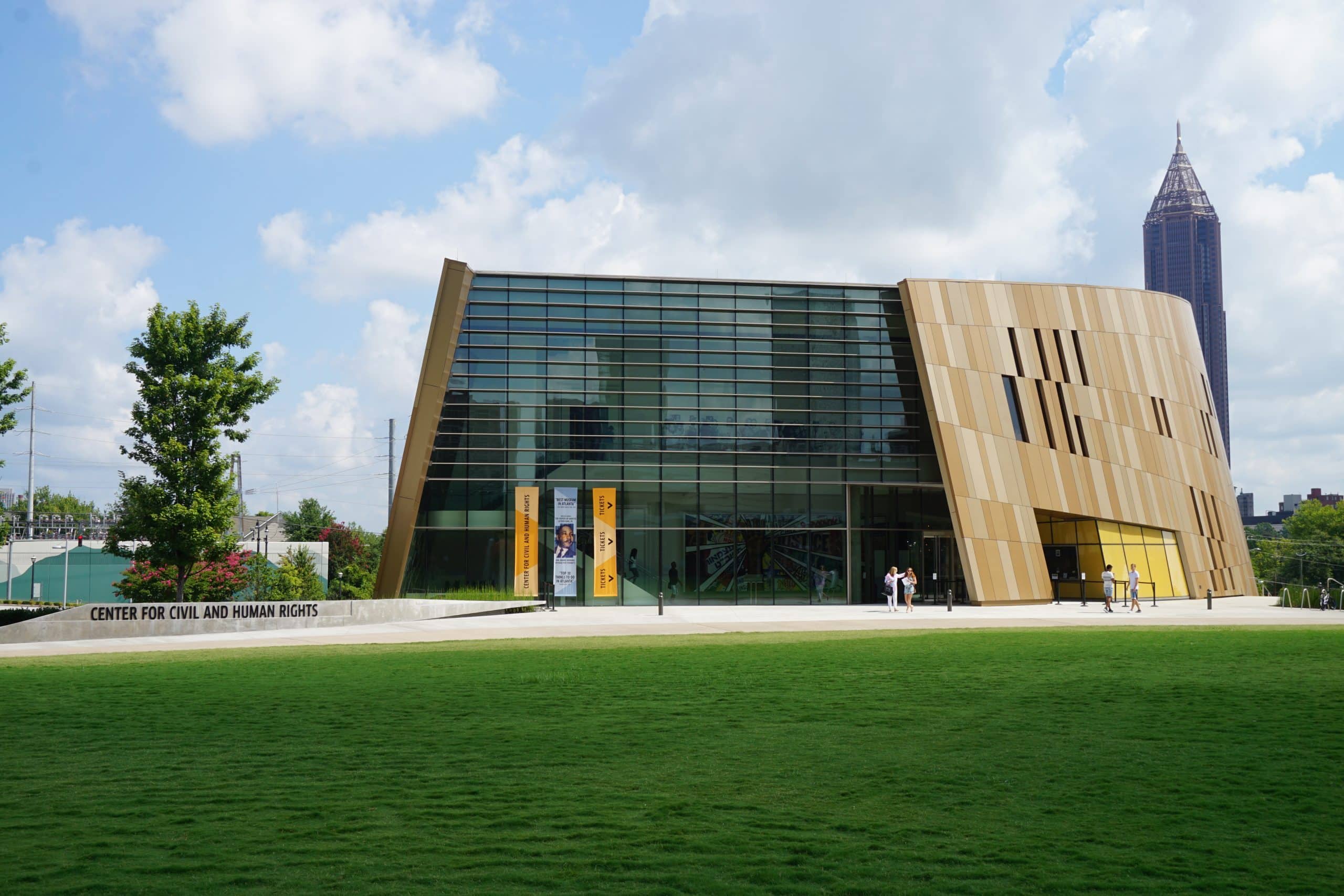 A modern building with large glass windows and tan angled panels, labeled "Center for Civil and Human Rights," sits next to a green lawn with a city skyscraper visible in the background under a partly cloudy sky.