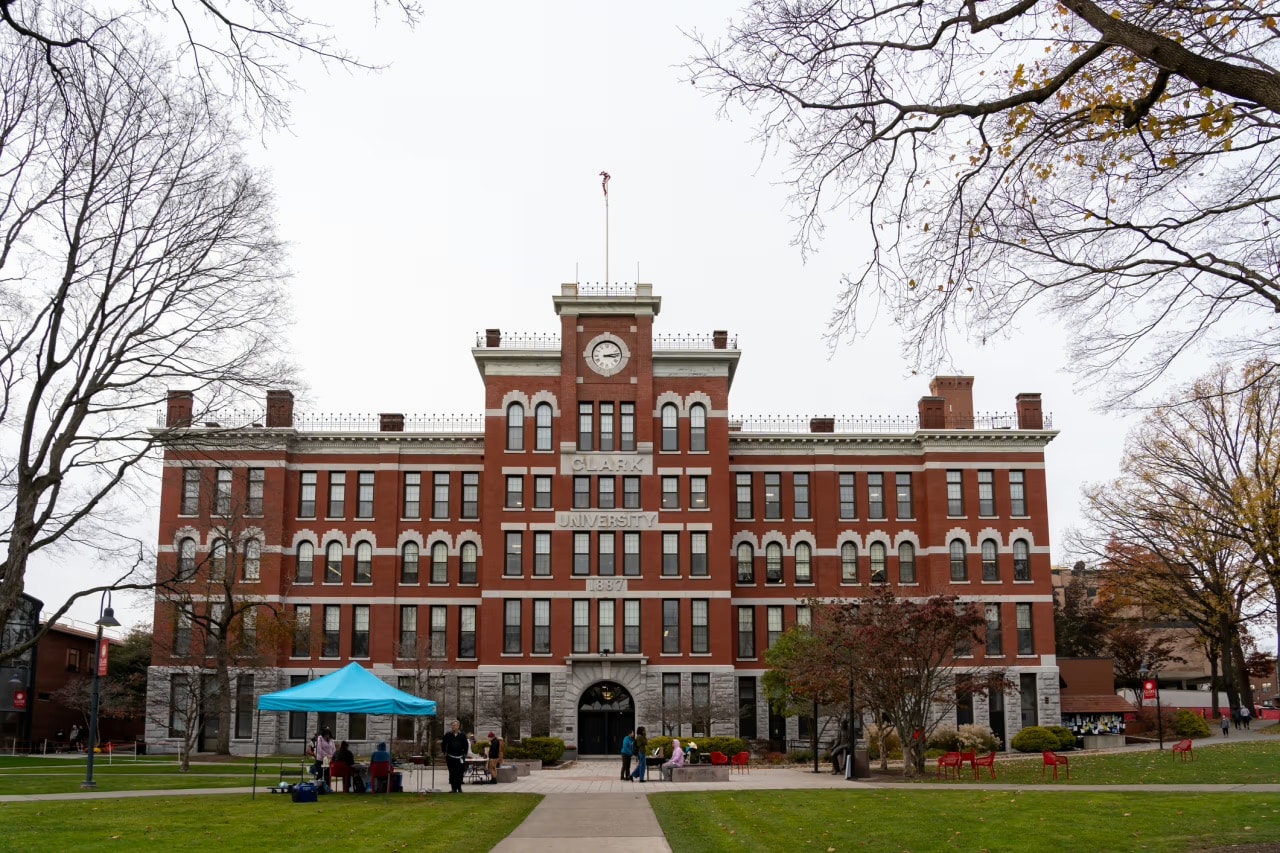 A large red-brick university building with a central clock tower and arched entrance, surrounded by bare trees and people near a blue tent on the lawn.
