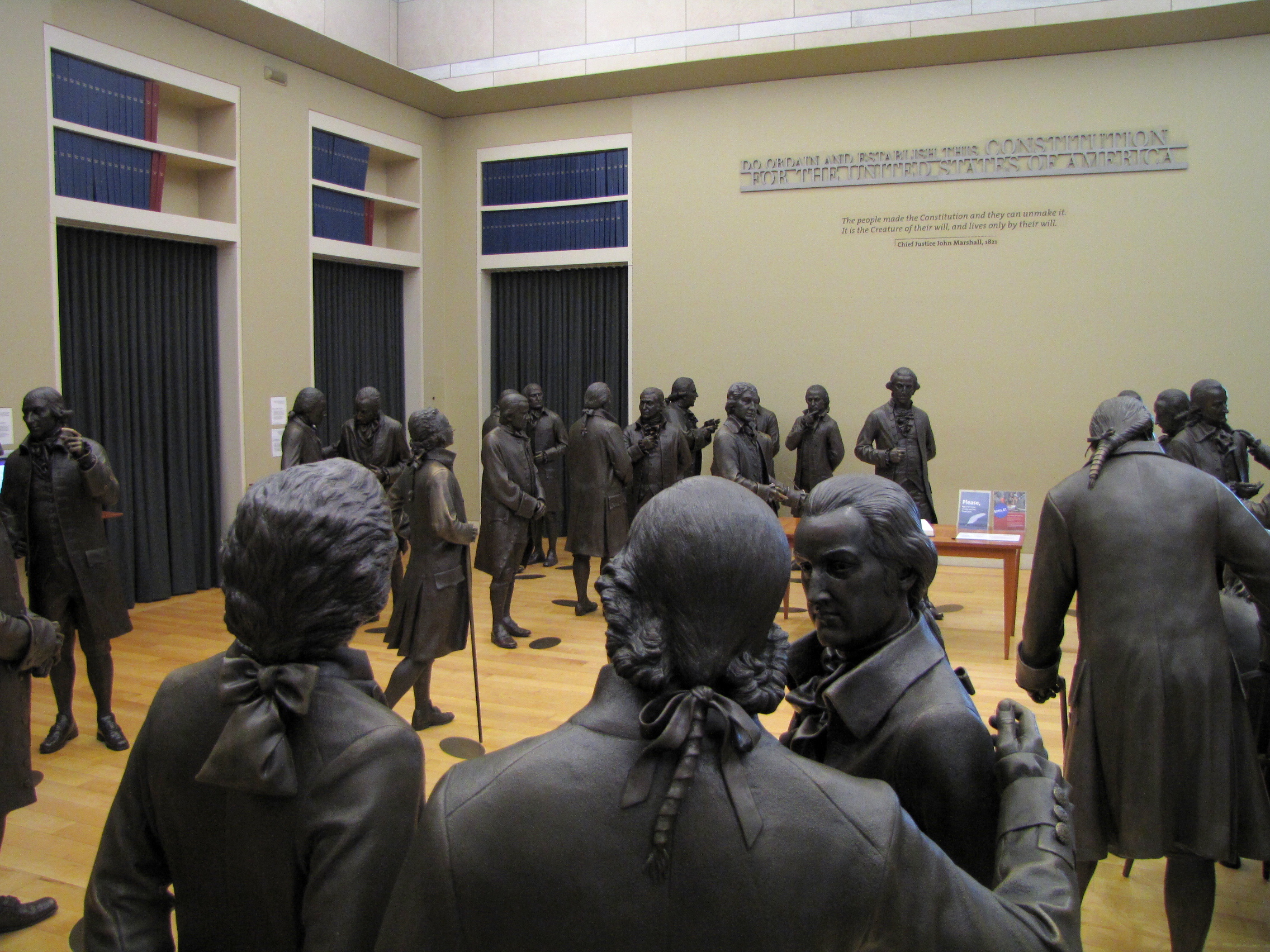 Lifelike bronze statues of historical figures in 18th-century attire stand in a museum exhibit at the National Constitution Center Philadelphia, gathered as if in discussion, with bookshelves and text about the U.S. Constitution behind them.