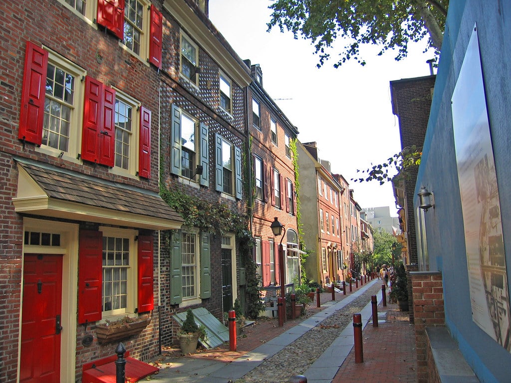 A narrow, historic street lined with brick houses featuring bright red shutters, green vines climbing the facades, and a cobblestone path, with trees and lampposts along the sidewalk on a sunny day.