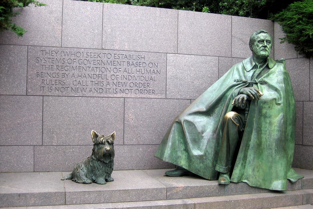 A bronze statue of Franklin D. Roosevelt seated in a cape beside his dog, Fala, with an engraved quote about government on a stone wall behind them at the FDR Memorial—an inspiring stop for Educational Field Trips to Washington DC.