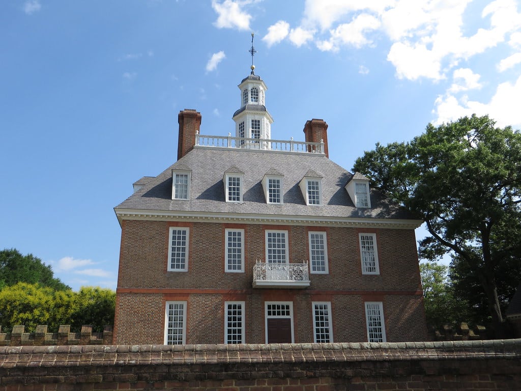 A large, symmetrical brick colonial-style building with tall chimneys, white-framed windows, a balcony, and a decorative cupola evokes the charm of a Colonial Williamsburg field trip, set against a blue sky with clouds and surrounded by trees.