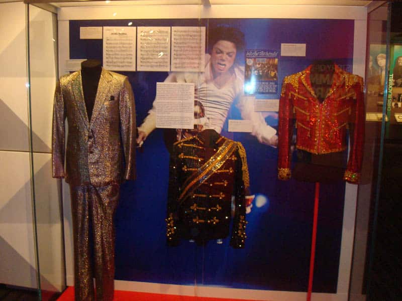 Three glittery costumes, including gold and red jackets, are displayed in a glass case with a photo of Michael Jackson performing in the background, along with sheet music and informational signs—a highlight for anyone on a 5 Day Los Angeles School Trip exploring the city's vibrant pop culture history.