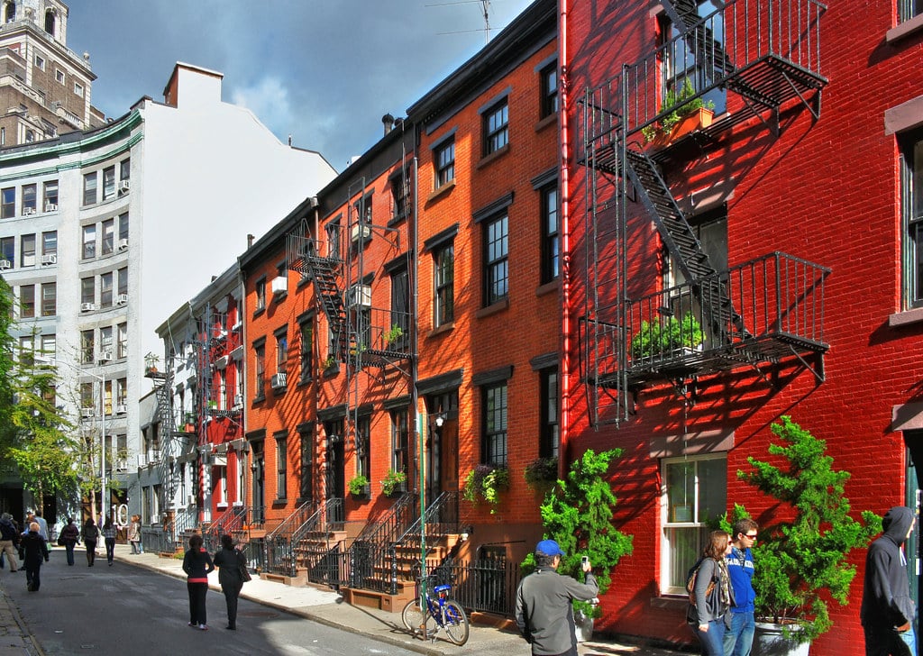 A sunny city street lined with red brick row houses, black fire escapes, and potted plants. People walk along the sidewalk, and trees and taller buildings are visible in the background.