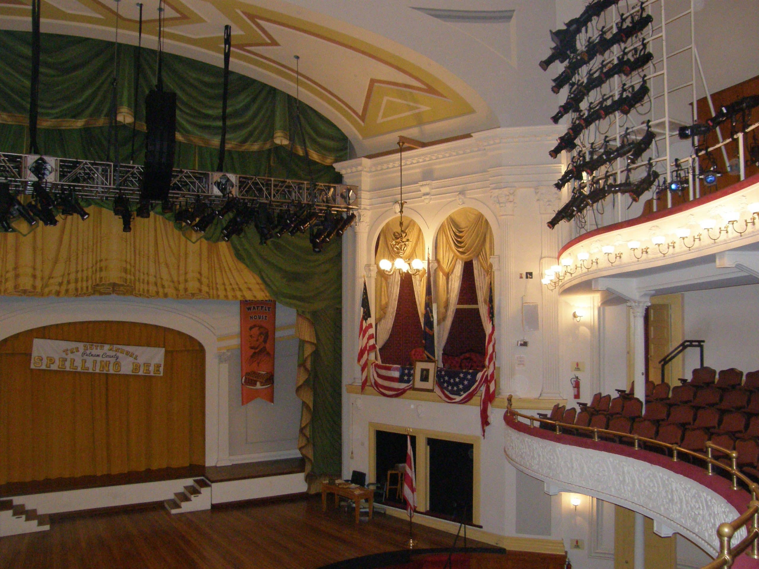 A historic theater interior with a wooden stage, yellow curtain, and green drapes—perfect for Educational Field Trips to Washington DC. American flags and patriotic bunting decorate the balcony above rows of red seats, with stage lighting rigs overhead.