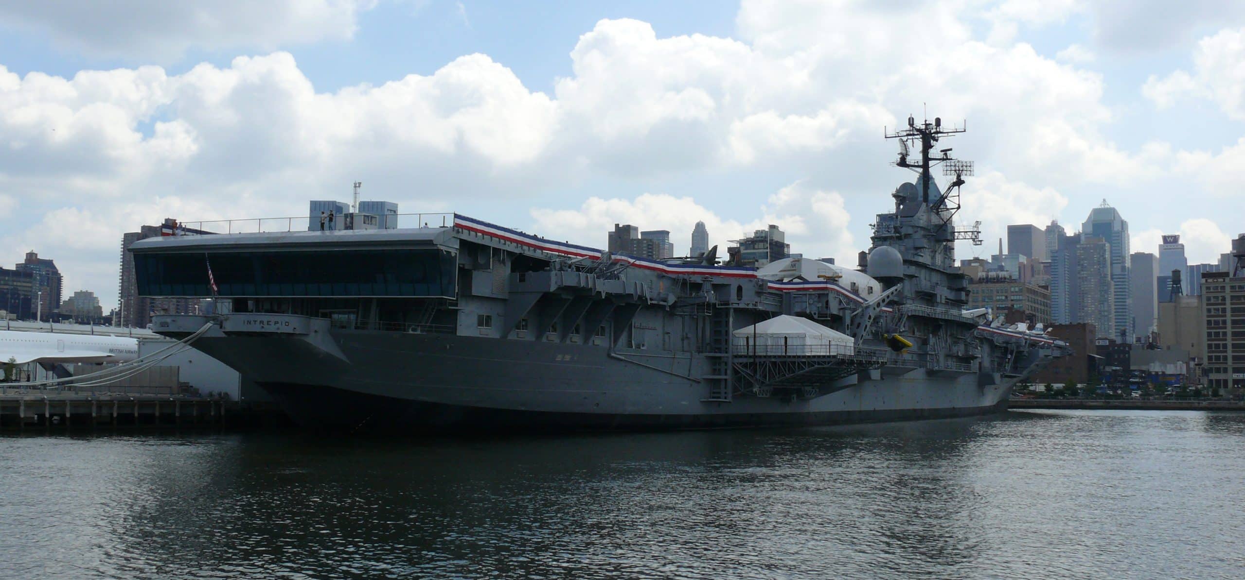 A large gray aircraft carrier, now one of the Best Museums in NYC for Educational Travel, is docked by the waterfront with city buildings and a cloudy sky in the background. The vessel features a prominent control tower and deck alongside the pier.