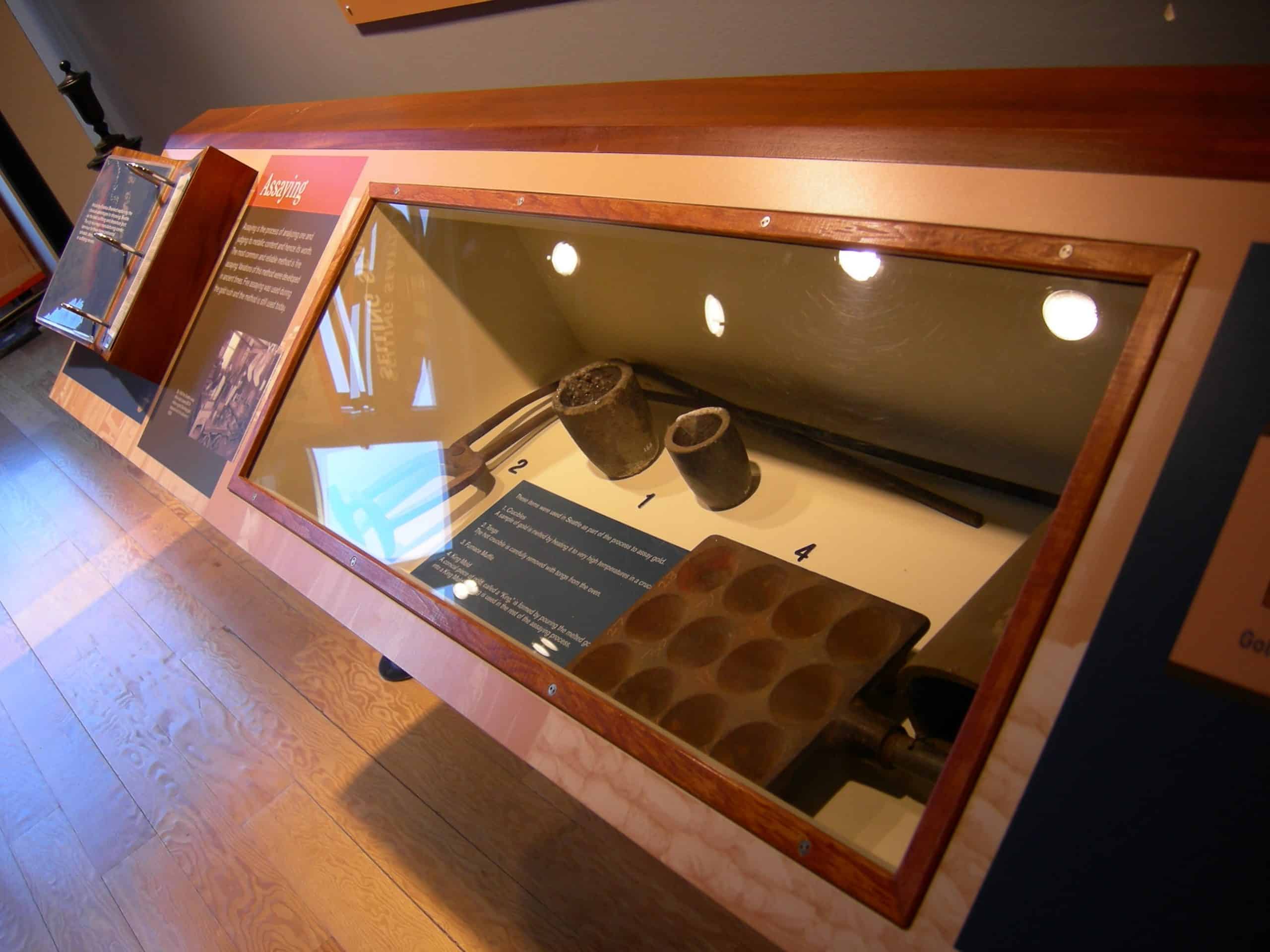 A museum display case—one of the Top 10 things to do on a Seattle Field Trip—contains historical logging tools, including two metal cauldrons, a long iron rod, and a rectangular iron mold with circular indentations, each labeled with a number.