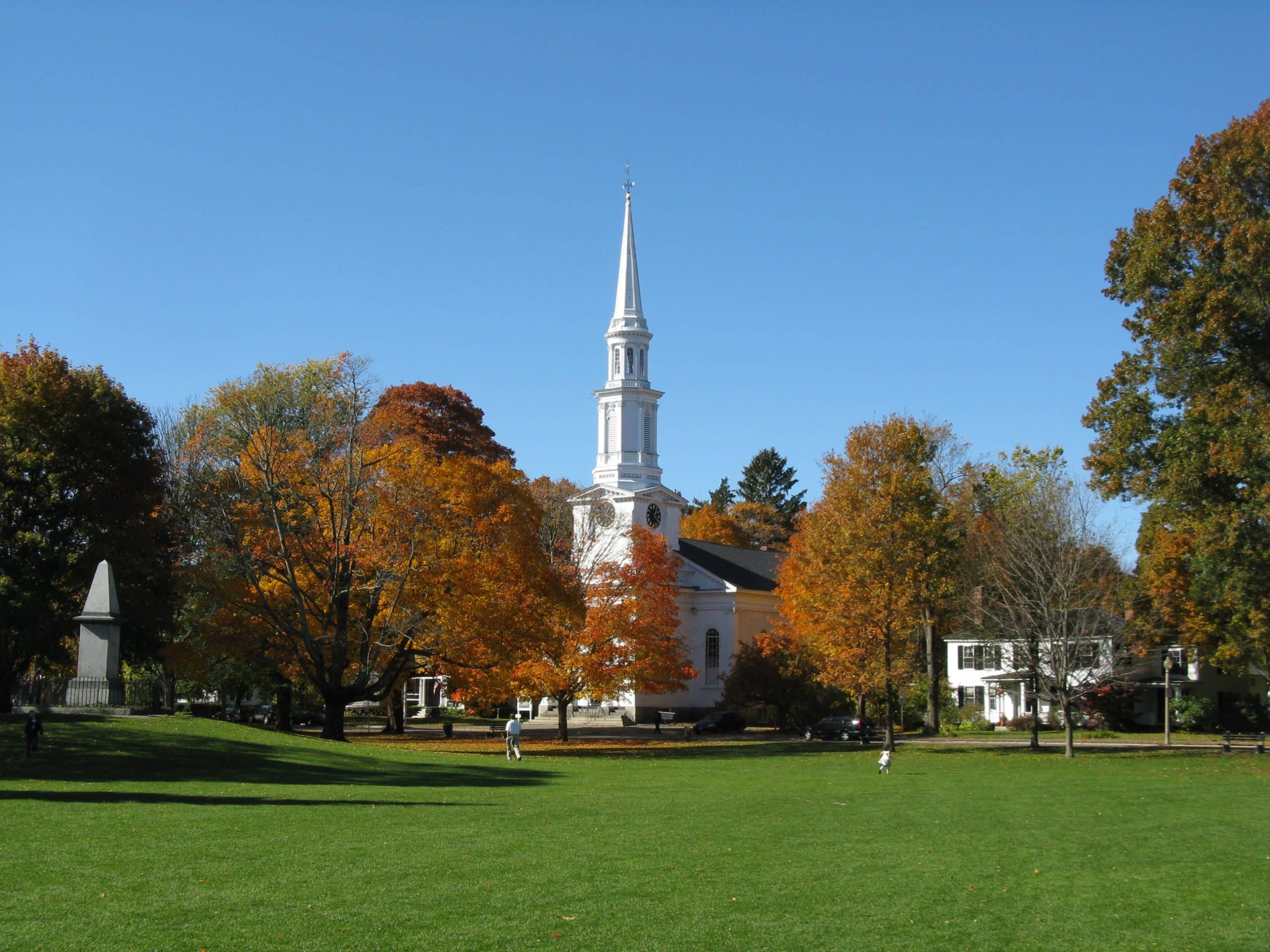 A white church with a tall steeple stands among colorful autumn trees, facing a green lawn under a clear blue sky. A few people walk on the grass, and houses are visible in the background.