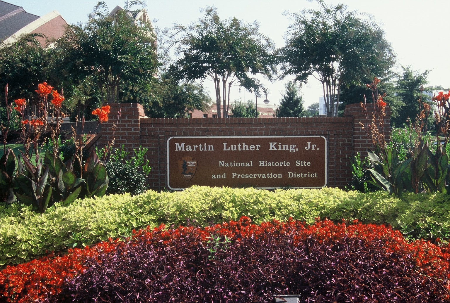 A brick sign reading "Martin Luther King, Jr. National Historic Site and Preservation District" stands surrounded by colorful flowers—an inspiring stop on your 3 Day Atlanta Field Trip—framed by lush greenery, trees, and a nearby building.