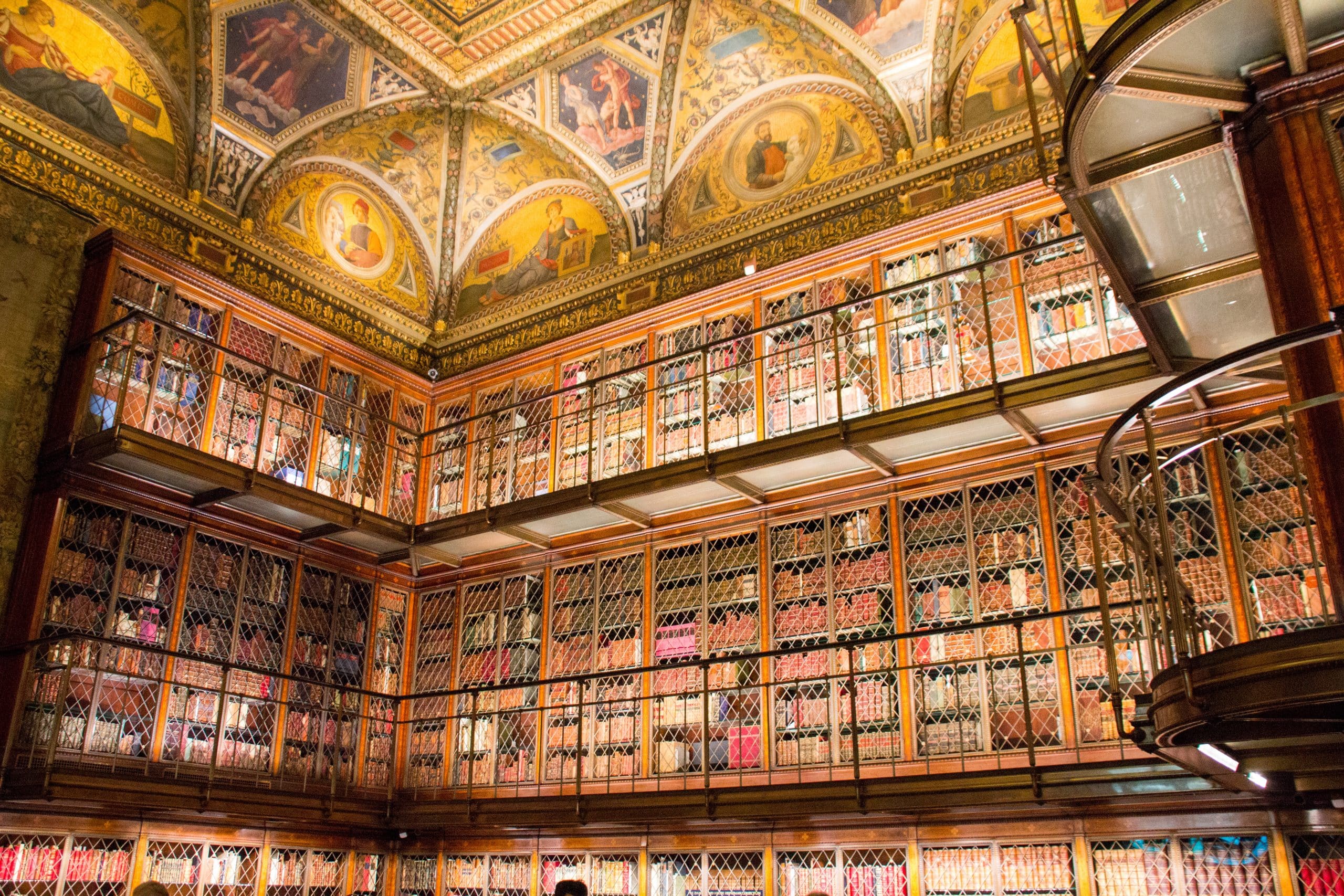 A grand library interior, reminiscent of the Best Museums in NYC for Educational Travel, features ornate painted ceilings and two levels of bookshelves behind decorative metal grates. Warm lighting highlights the intricate architecture.