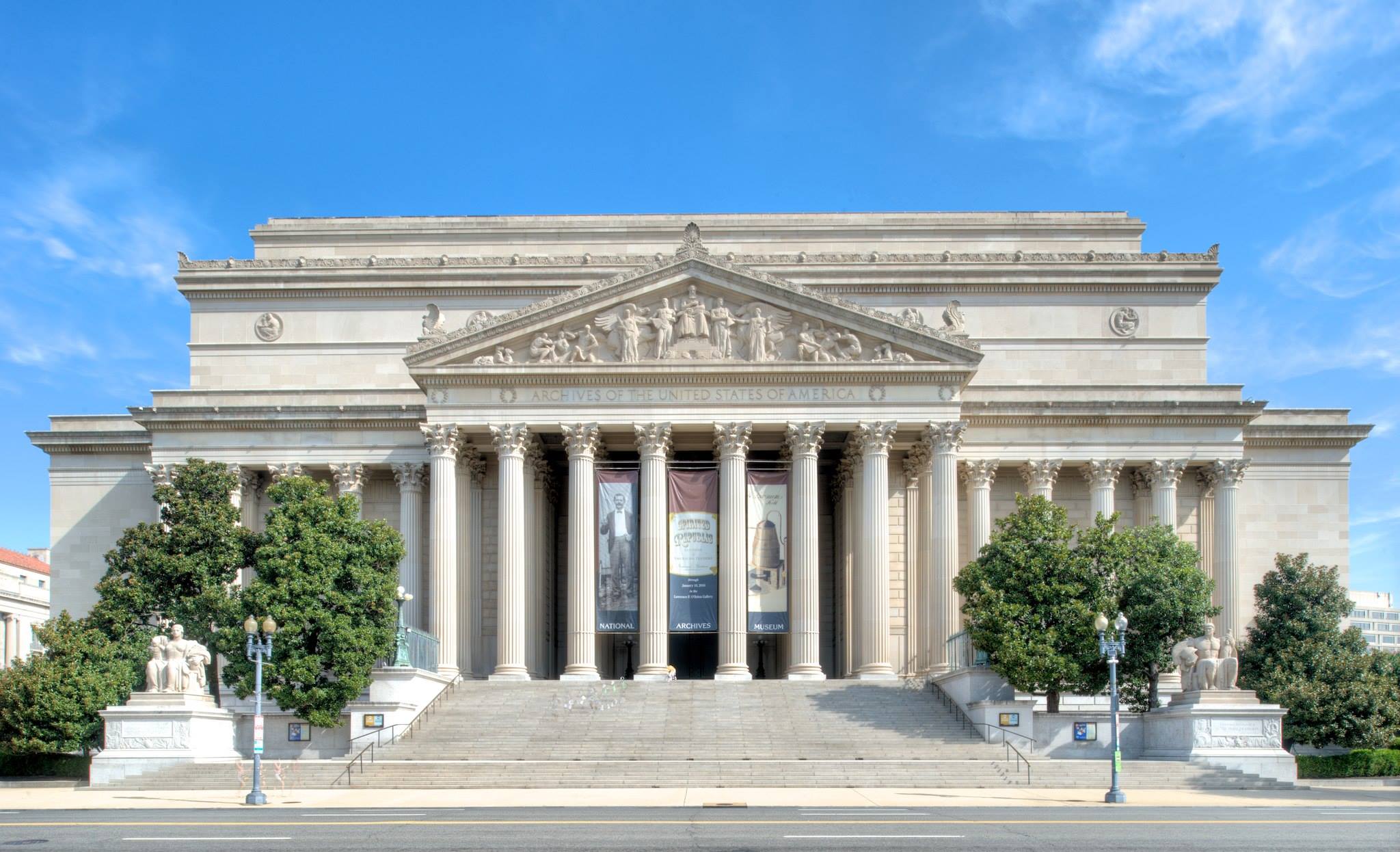 A neoclassical building with tall columns, large steps, and sculpted figures on the pediment—an ideal stop for educational field trips to Washington DC—flanked by trees and lampposts under a blue sky.