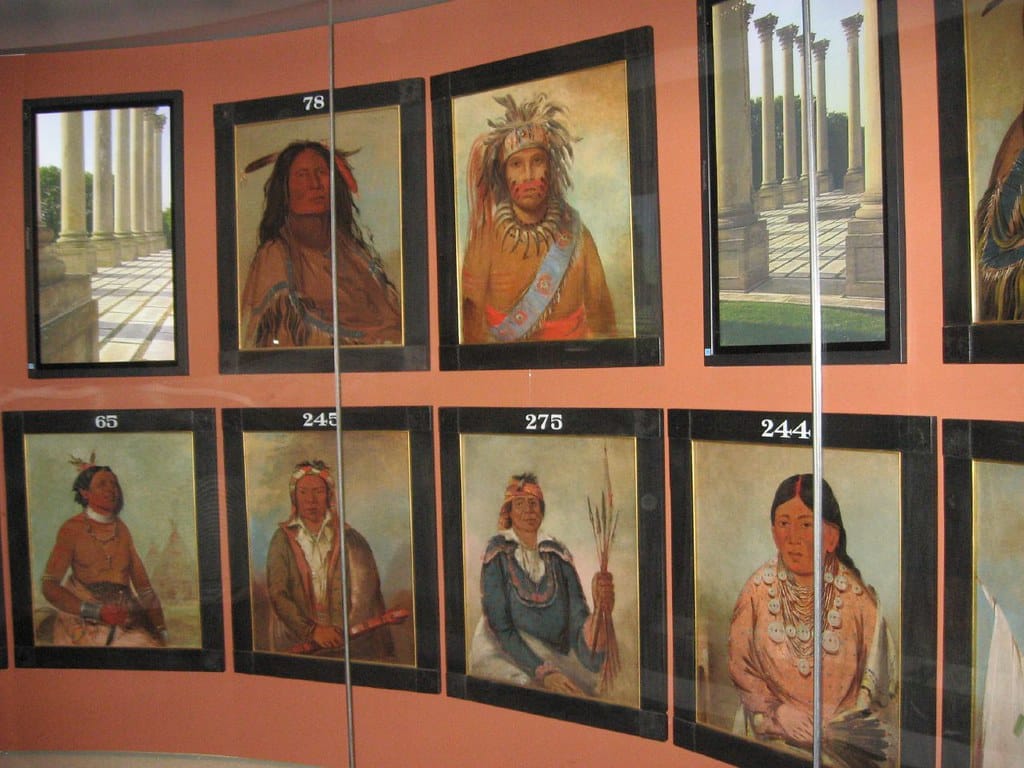 Paintings of Indigenous people in traditional attire are displayed on a curved wall, each labeled with a number—a unique visual highlight for STEM Washington DC school trip ideas. A reflection shows columns from another artwork or structure nearby.