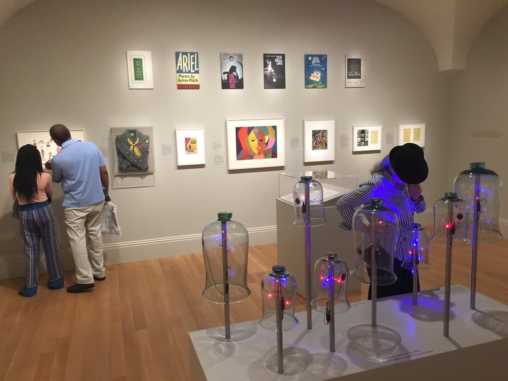 Two people view framed art on a gallery wall, while another examines glass sculptures glowing with blue and red lights. With wooden floors and white walls, this gallery is an inspiring stop for STEM Washington DC school trip ideas.