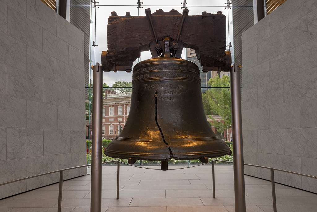 The Liberty Bell, one of the most iconic Philadelphia historical sites for students, is displayed indoors with its famous crack visible. A window behind it reveals a historic building and green trees outside.