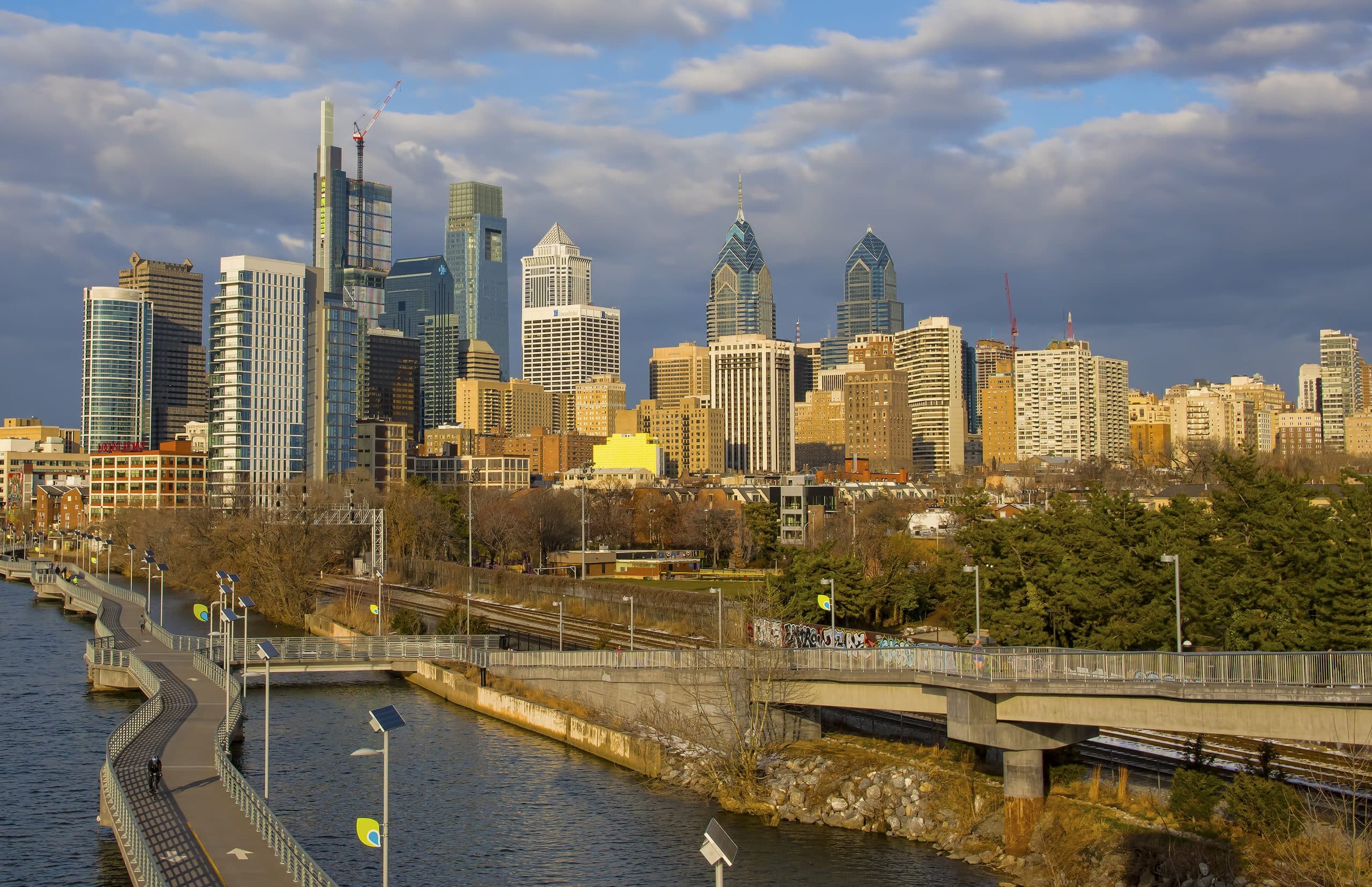 Philadelphia skyline with modern skyscrapers and historic buildings, seen from across a river with a curving pedestrian pathway and trees in the foreground under a partly cloudy sky.