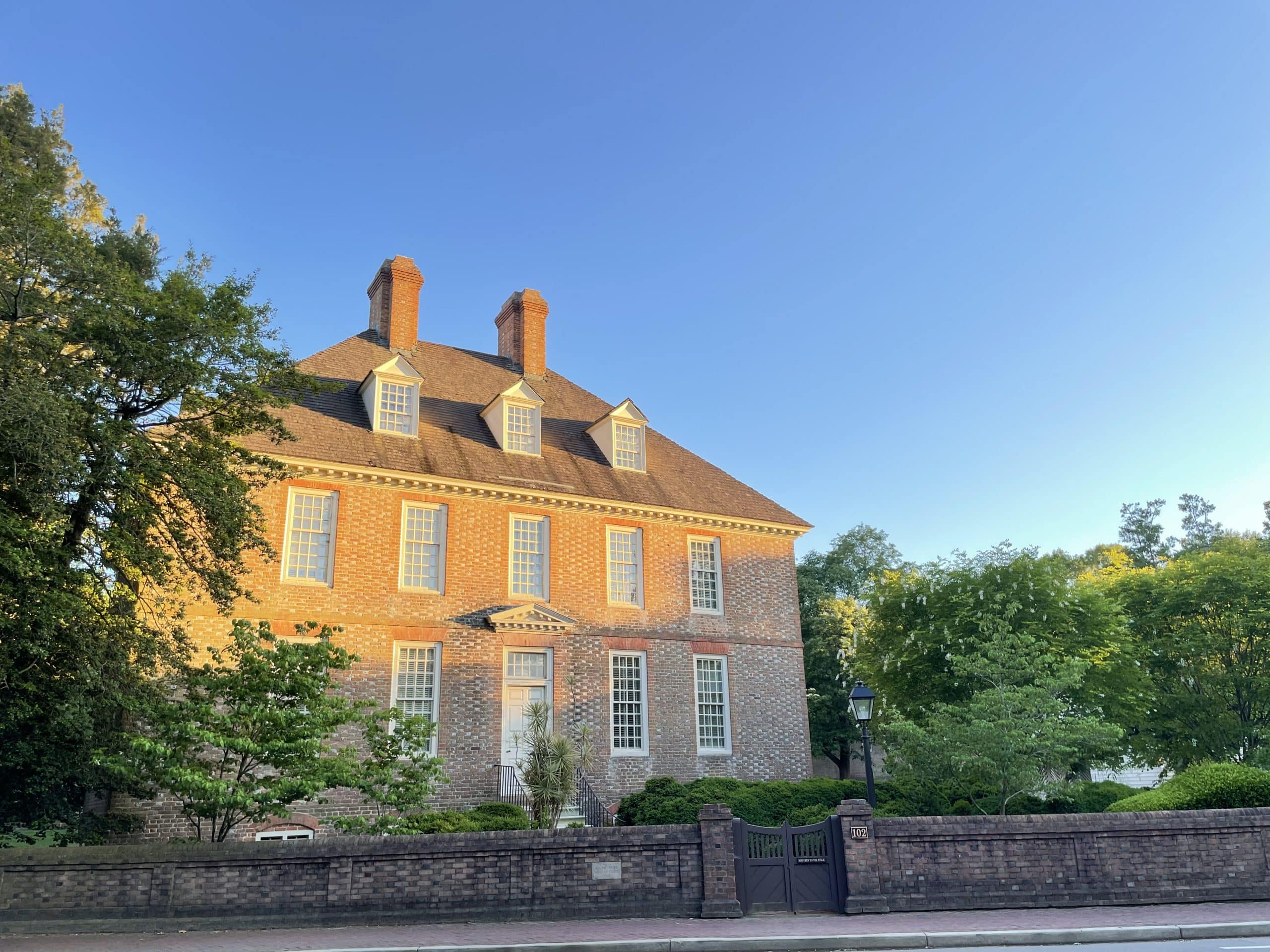 A large, historic brick house with tall chimneys and multiple windows is surrounded by trees and greenery under a clear blue sky—an ideal scene for a Williamsburg School Trip. A brick fence with a gate stands in front of the house.