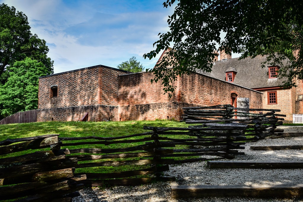 A historic brick building with red window frames, often seen on a Colonial Williamsburg field trip, is surrounded by a rustic wooden split-rail fence and trees under a partly cloudy sky. Gravel steps lead up to the fenced area.