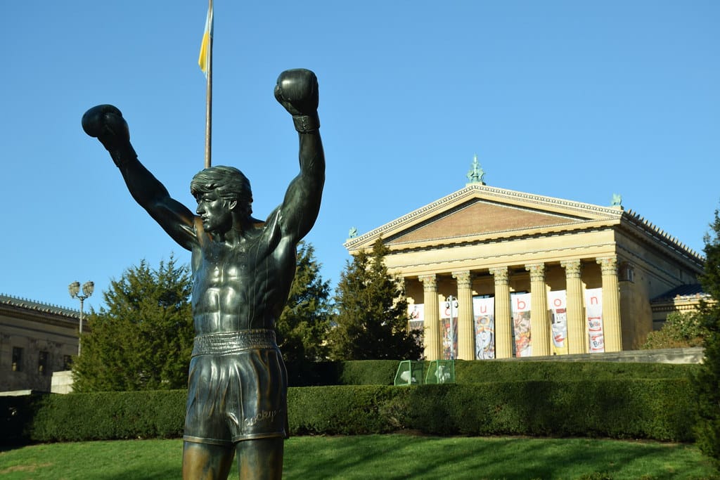 A bronze statue of a boxer with arms raised stands in front of the Philadelphia Museum of Art, with a clear blue sky and a flag visible in the background.