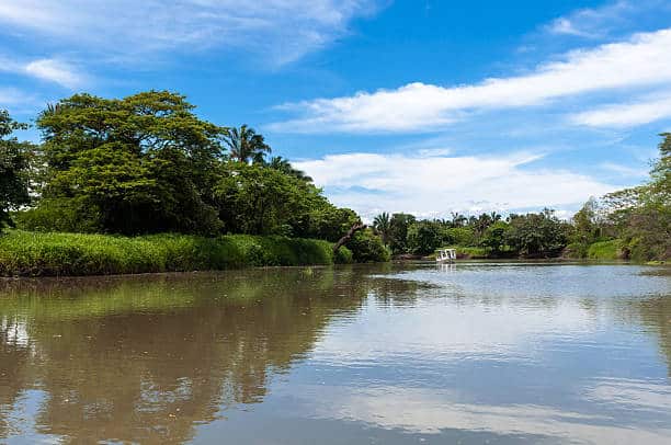 A calm river reflecting green trees and blue sky with scattered white clouds, surrounded by lush vegetation; a small white structure is visible in the distance along the riverbank.