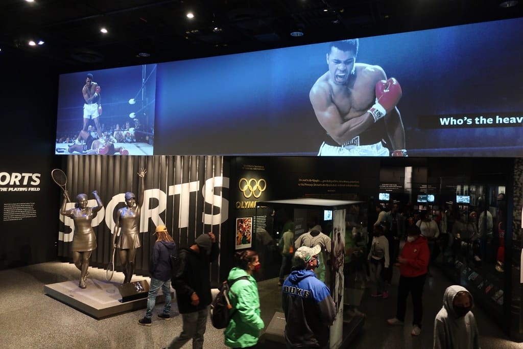 This museum exhibit, highlighted among the Best Smithsonian Museums for Student Groups, features sports displays, mannequins in athletic poses, and a striking image of a boxer mid-punch as visitors explore under dim lighting.