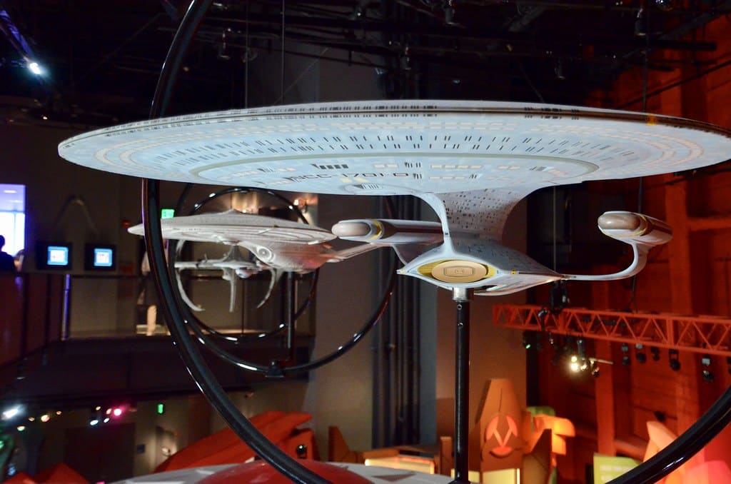 A detailed model of the Starship Enterprise from Star Trek is suspended from the ceiling in a museum exhibit, part of a Seattle School Trip, with another starship visible in the background under dramatic lighting.