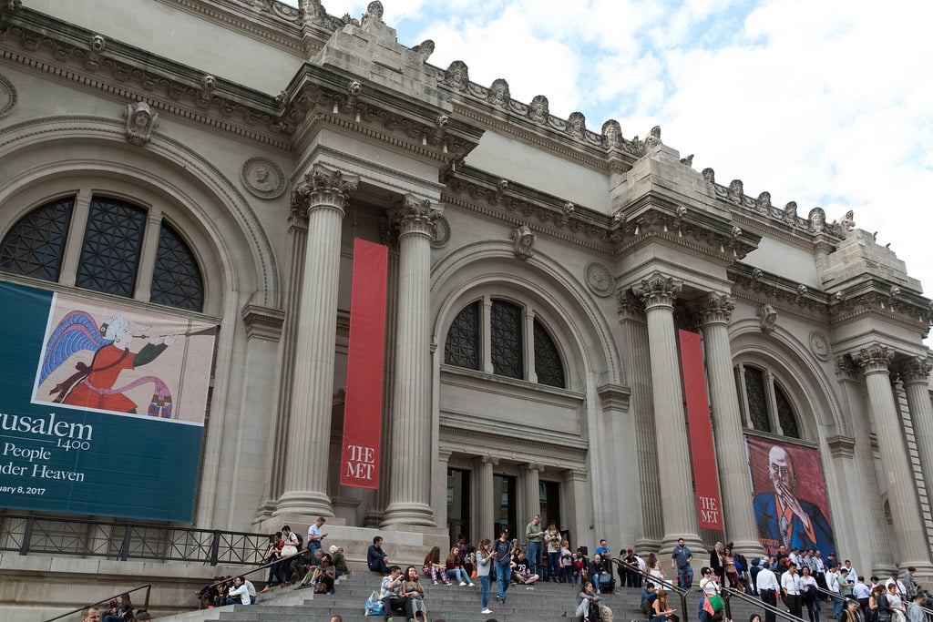 The front entrance of The Met museum, one of the best museums in NYC for educational travel, features large columns, banners, and many people sitting and walking on the wide stone steps under a partly cloudy sky.