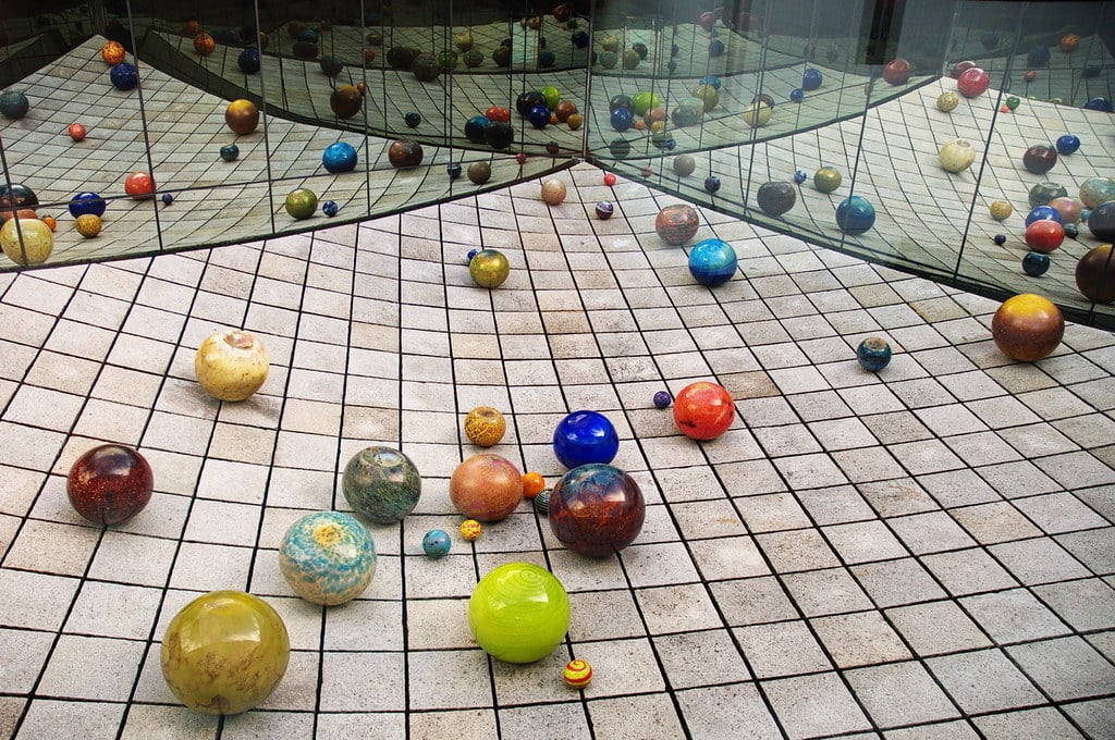 Glass spheres of various sizes and colors rest on a tiled grid floor, with mirrored walls reflecting both the spheres and the grid—like an endless wonder you might find on a Seattle School Trip.