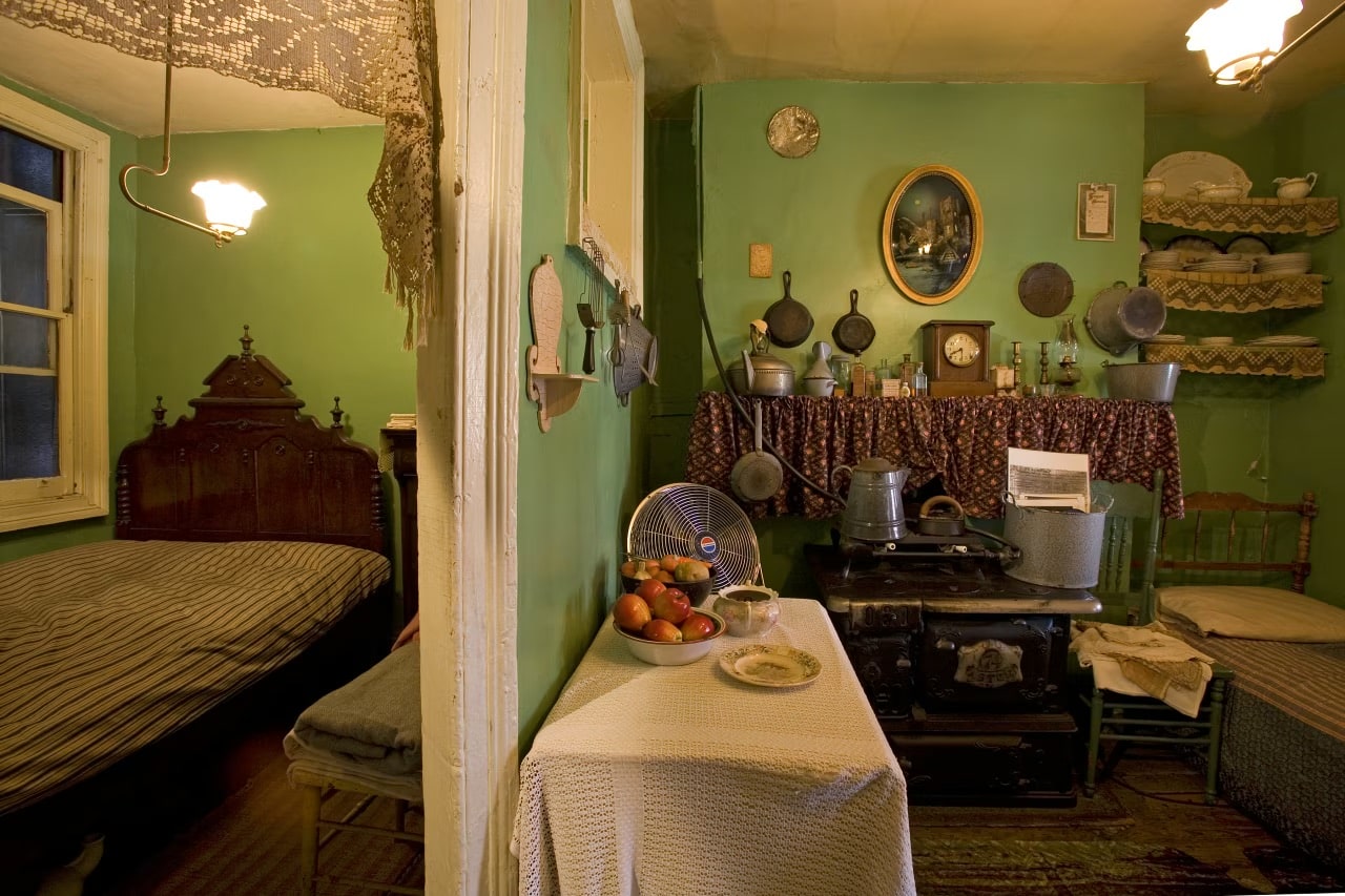 A cozy vintage kitchen with green walls, a table set with fruit and a plate, an old stove, and shelves with dishes evokes the charm found in exhibits at the Best Museums in NYC for Educational Travel. Warm lighting gives the space a nostalgic feel.