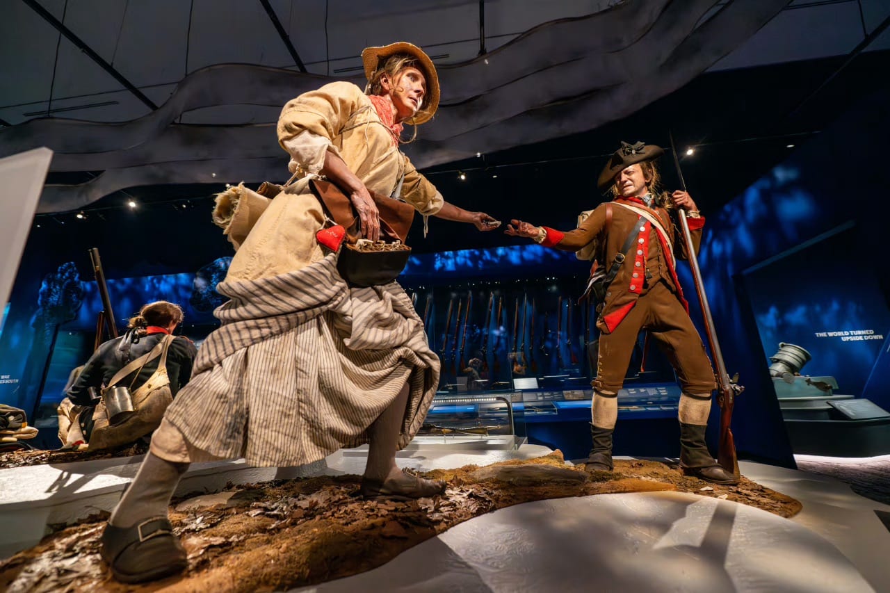 Museum display of two realistic mannequins: a woman in colonial attire hands an object to a Revolutionary War soldier in uniform, with historical artifacts in the background. This engaging exhibit provides an immersive glimpse into American history, making it an excellent addition to any tour of Philadelphia Historical Sites for Students.