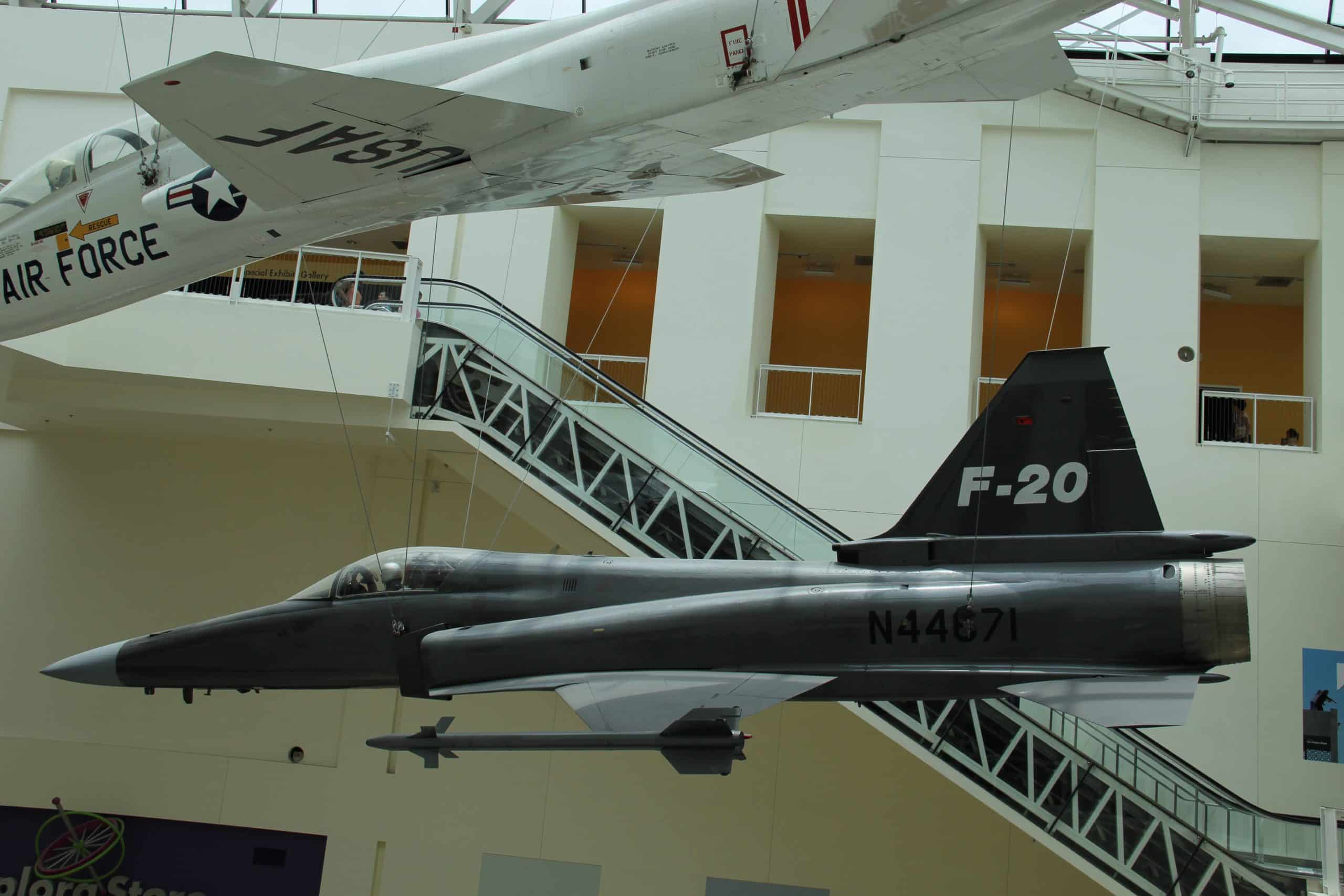 Two aircraft are suspended in a museum; the foreground jet is labeled "F-20 N44671," and behind it is a white jet marked "USAF Air Force." Both are displayed indoors near railings and a glass ceiling, making this an exciting highlight for students on a 5 Day Los Angeles School Trip.