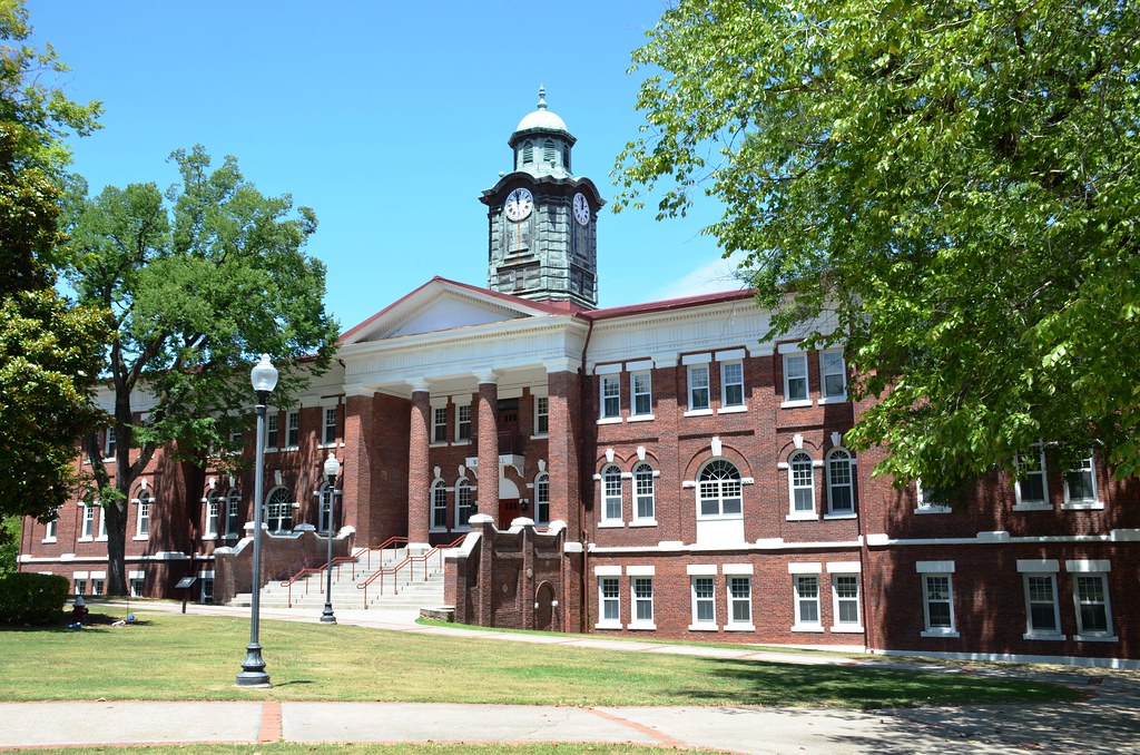 A historic red-brick building with white columns, large windows, and a central clock tower stands behind a grassy lawn, shaded by trees under a clear blue sky.