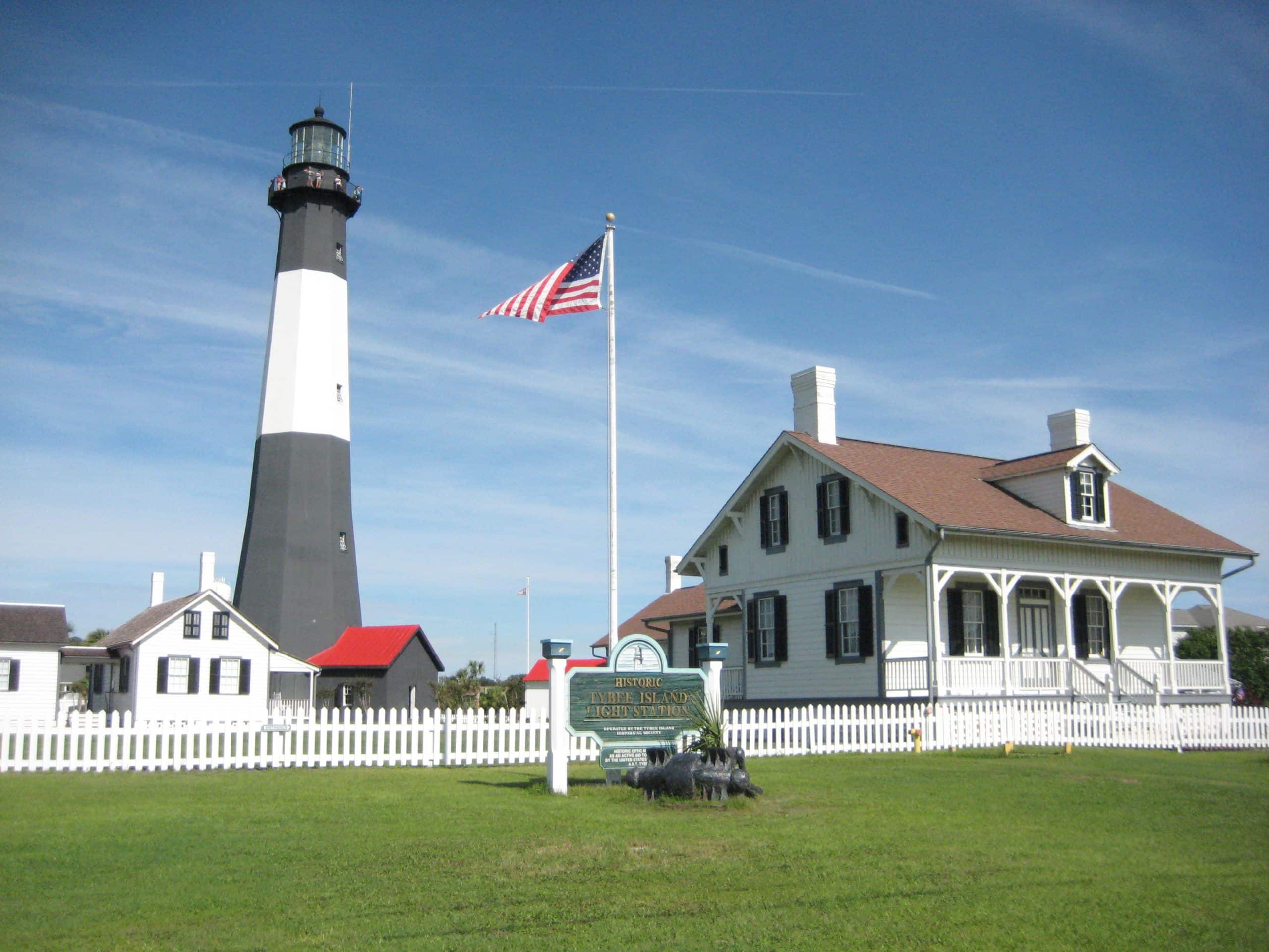 A tall black and white lighthouse stands next to a white house with a porch, white picket fence, and an American flag waving in front under a clear blue sky—a perfect Field Trip Savannah GA destination.