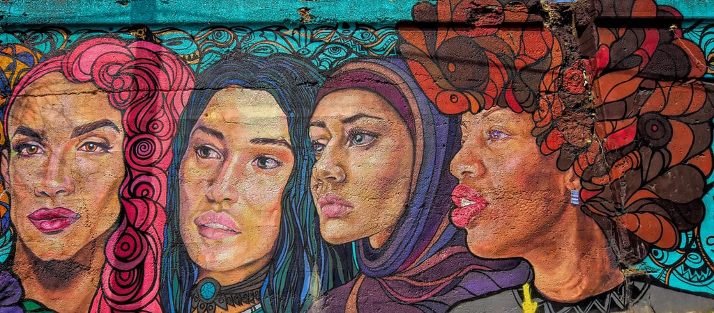 A colorful mural depicts four diverse women with unique hairstyles and accessories, including a headscarf and large earrings, reflecting global influences and student travel trends for 2027 against a vibrant turquoise background with intricate patterns.