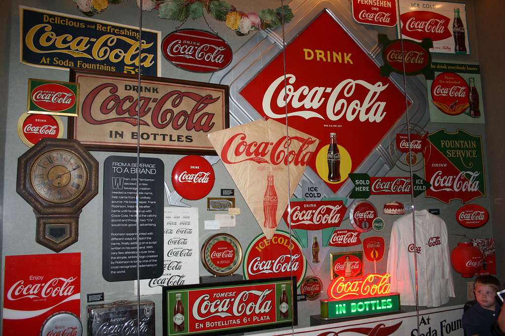 A wall covered with various vintage Coca-Cola signs, advertisements, clocks, and memorabilia—mostly in red, white, and green—creates a nostalgic scene. A young child explores in the corner during a 3 Day Atlanta Field Trip.