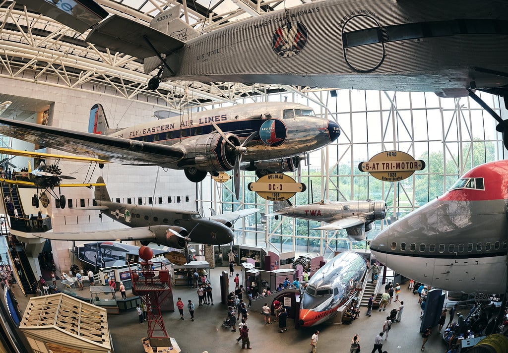 Several vintage airplanes, including a Boeing 747 nose, Ford Tri-Motor, and Douglas DC-3, are suspended from the ceiling in a spacious, sunlit museum—one of the Best Smithsonian Museums for Student Groups—filled with curious visitors.