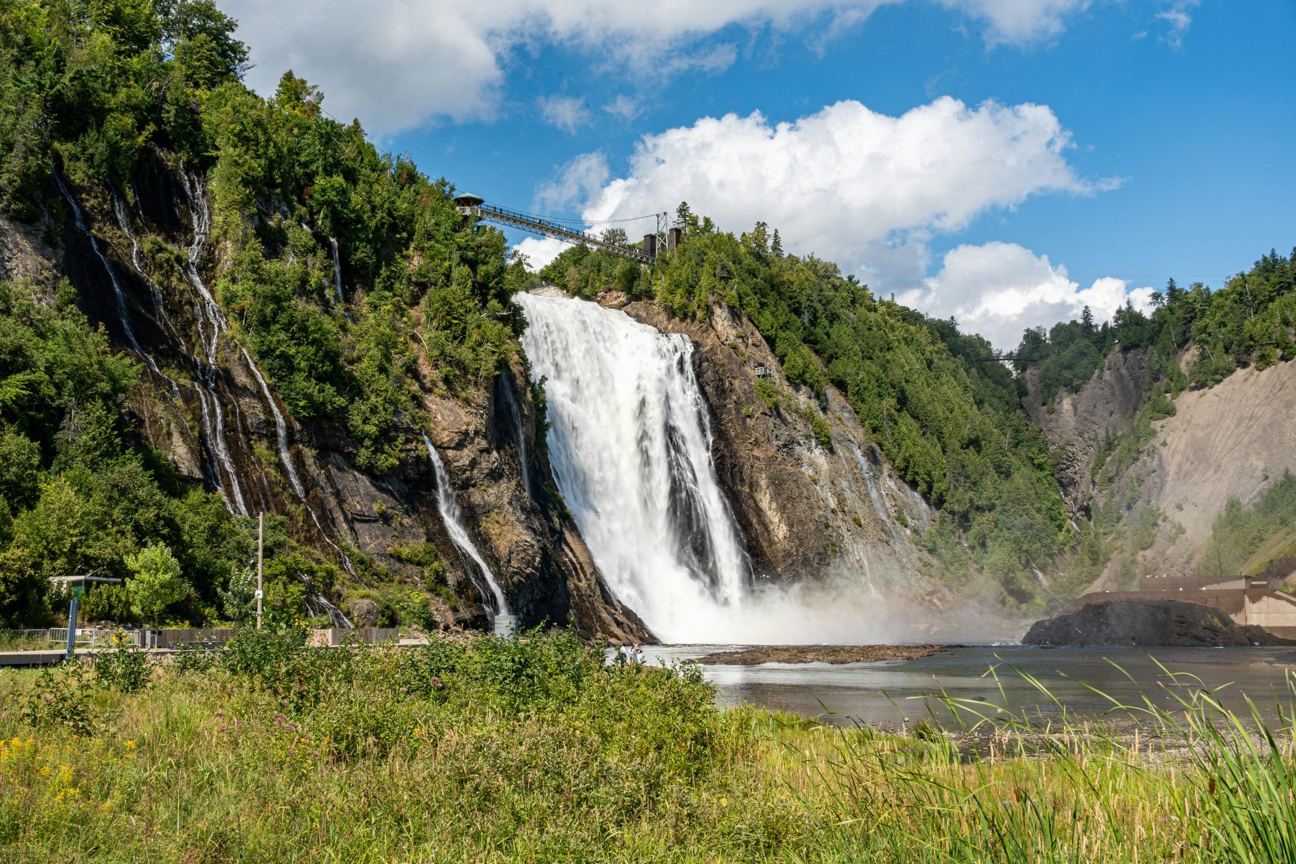 A large waterfall cascades down a rocky cliff surrounded by lush green trees, with a pedestrian bridge spanning above the falls—one of The Most Popular Student Travel Destinations for 2027—mist rising at the base under a blue sky with clouds.