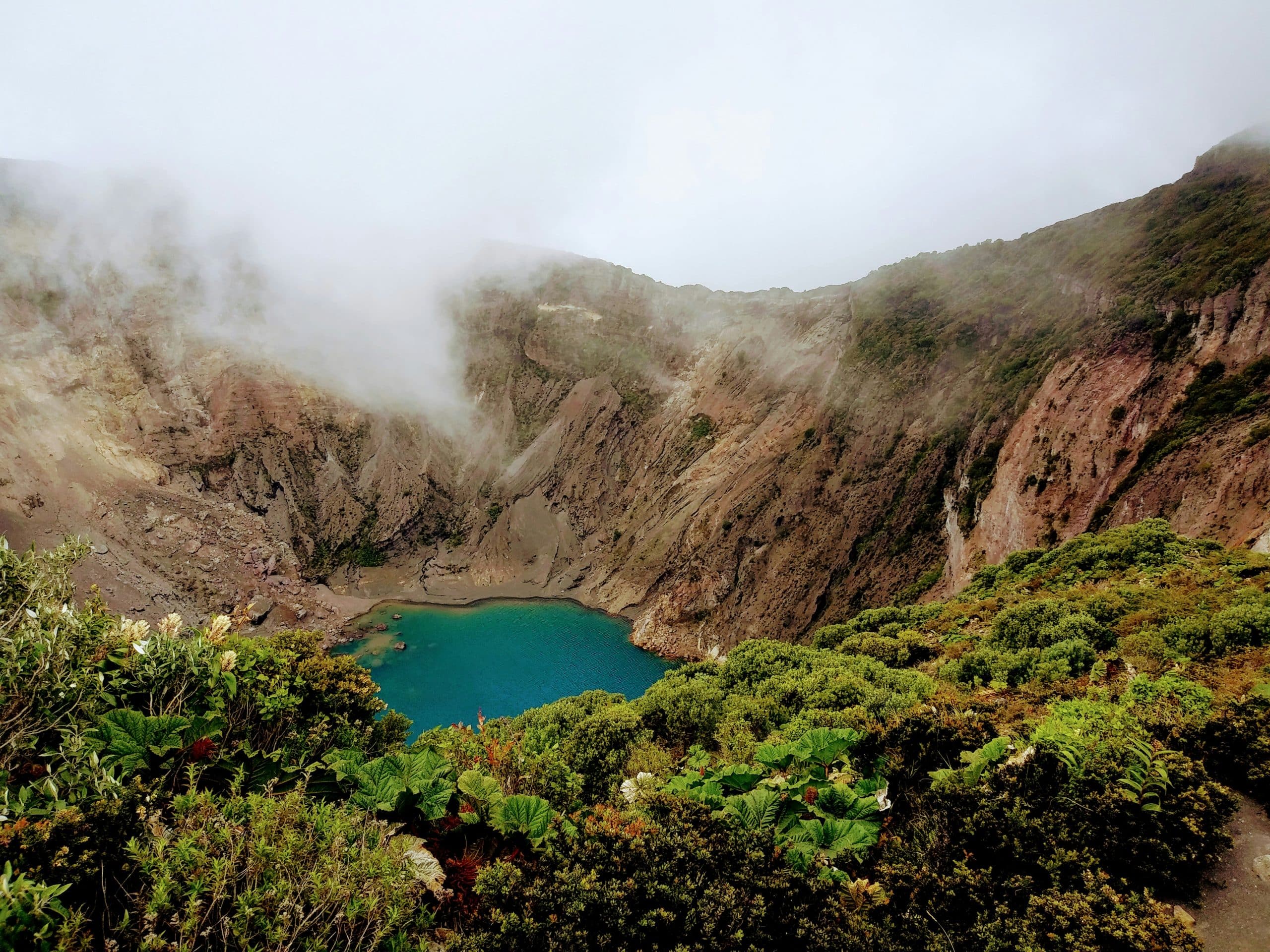 A turquoise crater lake surrounded by steep rocky cliffs and lush green vegetation, with mist and clouds partially covering the landscape.