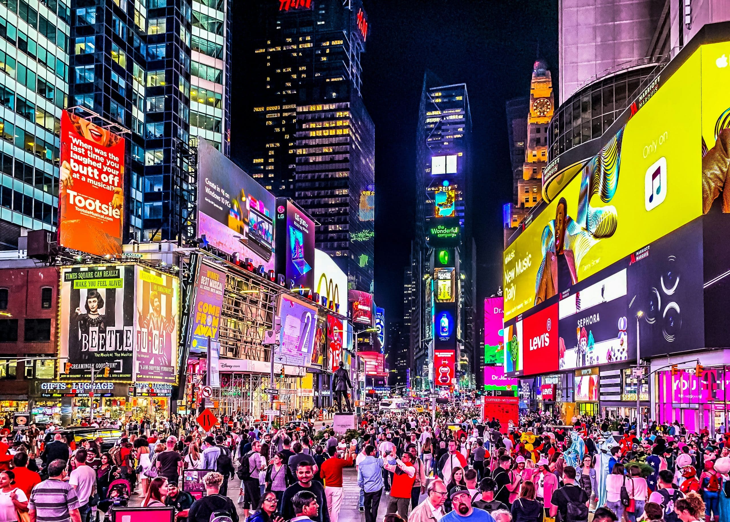 A large crowd gathers in Times Square at night, surrounded by bright, colorful digital billboards and advertisements lighting up the tall buildings on all sides.