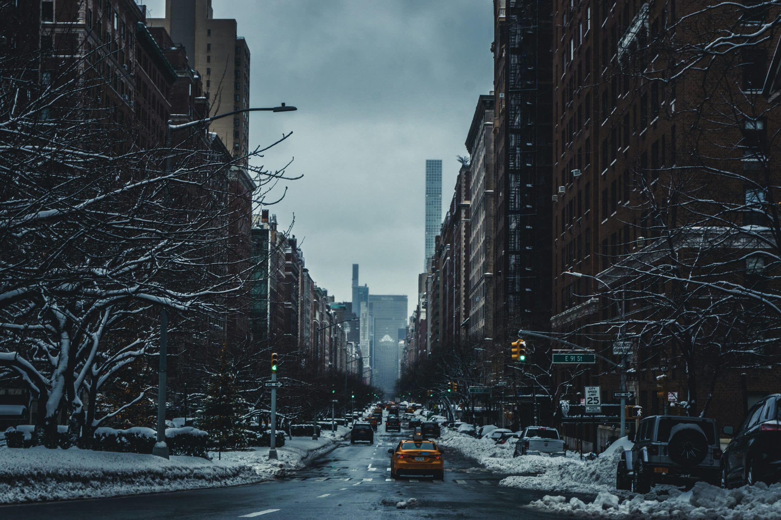 A snowy city street lined with tall buildings and bare trees, cars driving along wet pavement. A yellow taxi is in the center—echoing the urban vibrancy shaping Student Travel Trends for 2027 under a cloudy, gray sky.