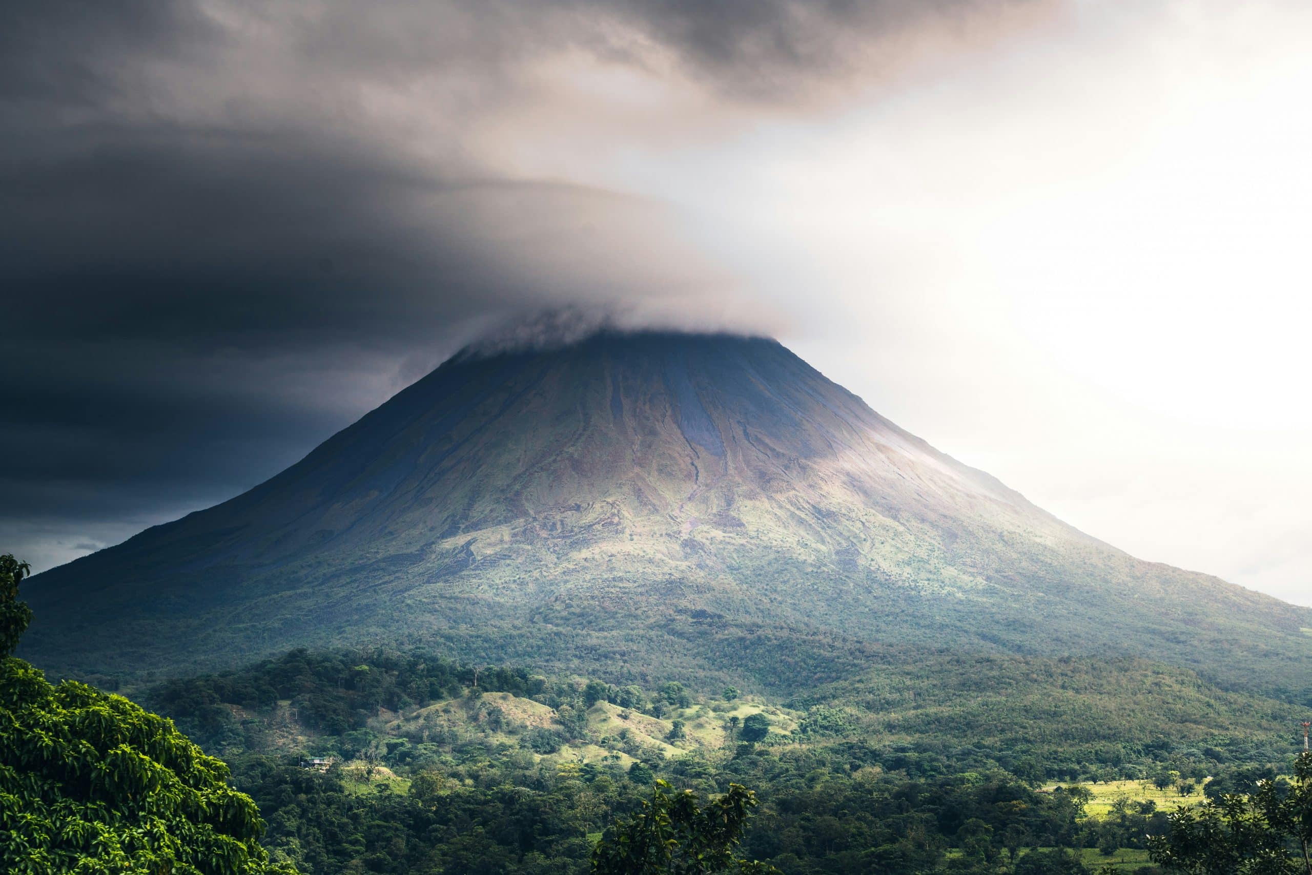A volcano with a cloud-covered summit rises above a lush green landscape, illuminated by sunlight breaking through the cloudy sky—a stunning scene among The Most Popular Student Travel Destinations for 2027.